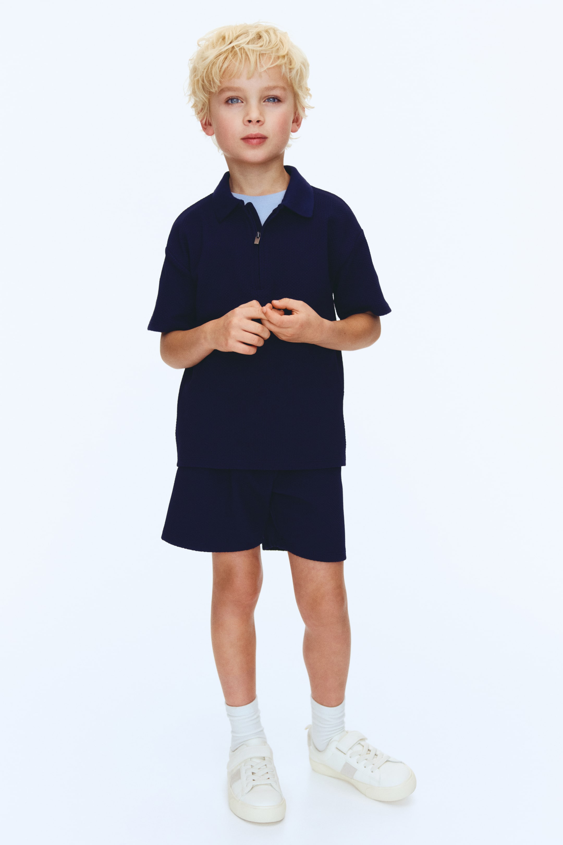 Προβολή μεγαλύτερης εικόνας: Ένα μπλε ναυτικό ριγέ πόλο μπλουζάκι με κοντά μανίκια και αντίστοιχο σορτς παρουσιάζονται από μπροστά. Το πόλο μπλουζάκι διαθέτει γιακά και μισό φερμουάρ, με ένα ανοιχτό μπλε μπλουζάκι να φαίνεται από κάτω, και το σορτς συντονίζεται στο χρώμα και στην υφή με ρίγες.