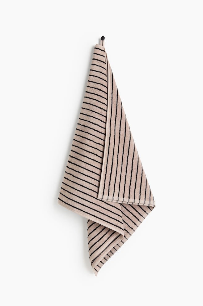 A bath towel is hanging from a black hook by a loop, presenting its front. It features beige terry fabric with black yarn-dyed stripes, appearing both horizontally and vertically due to its fold. - 1
