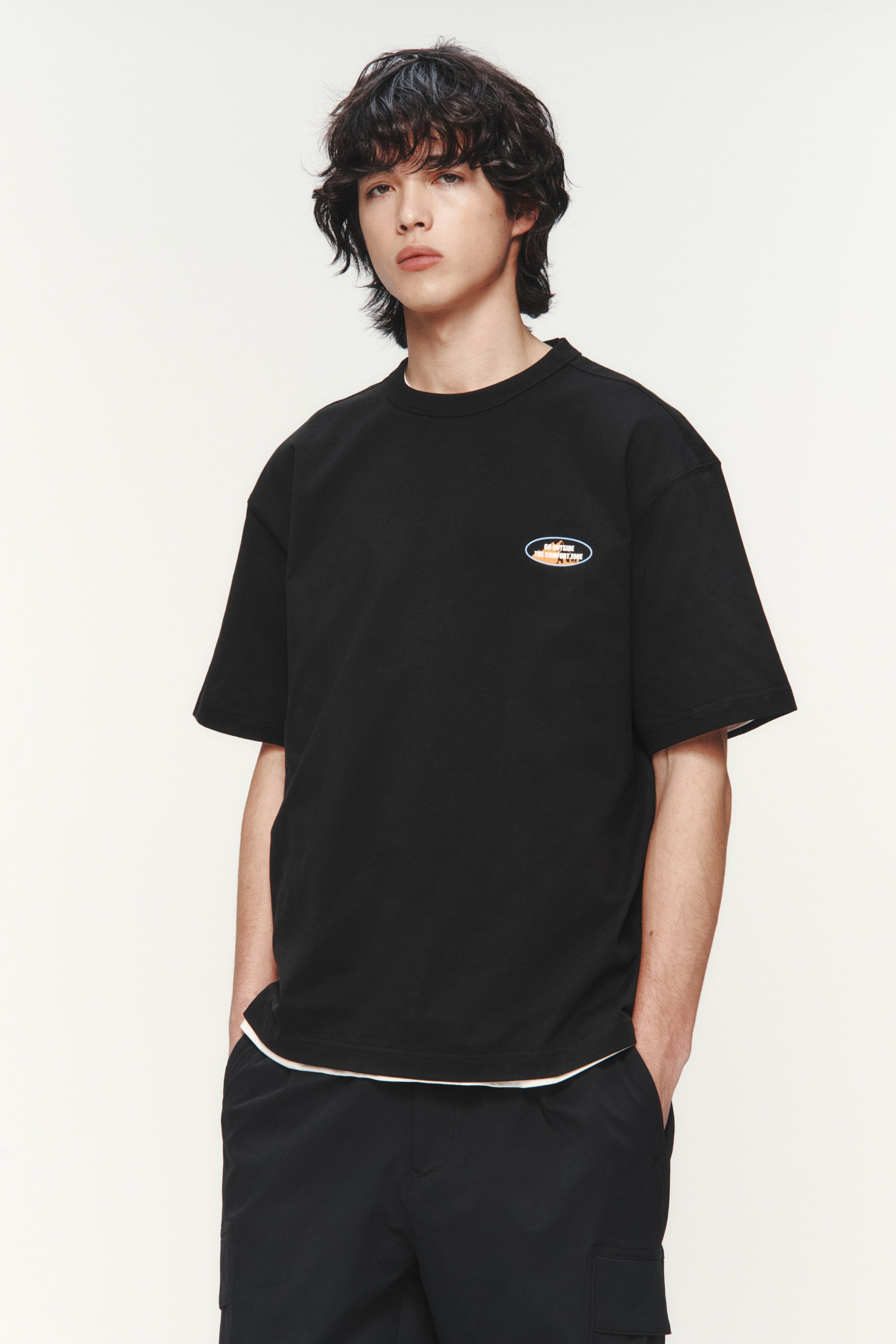 Xem ảnh lớn hơn: Áo thun cổ tròn màu đen được giới thiệu, nổi bật với họa tiết hình bầu dục ở phần ngực trái cùng chữ sáng và gradient màu cam, và tay áo ngắn, dáng rộng. Lớp áo trắng bên dưới lộ ra ở gấu áo.