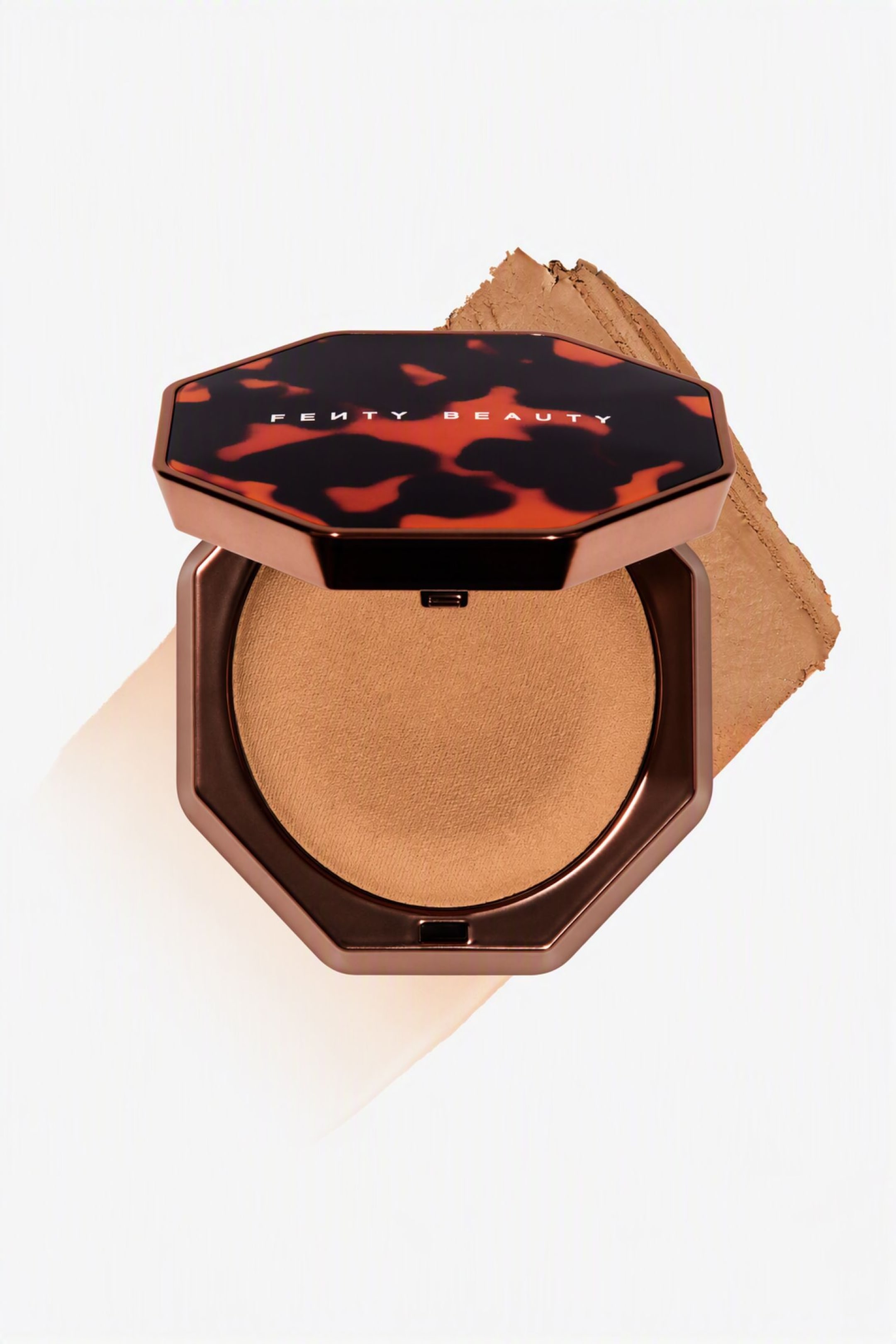 Visa större bild: En öppen, åttkantig Fenty Beauty-kompakt visar en jämn, bruntonad pressad bronzer i en rund panna. Den metalliskt bronsfärgade kompakten vilar på ett diagonalt penseldrag av produkten, med det upphöjda locket som visar ett sköldpaddsmönster och vit varumärkestext.