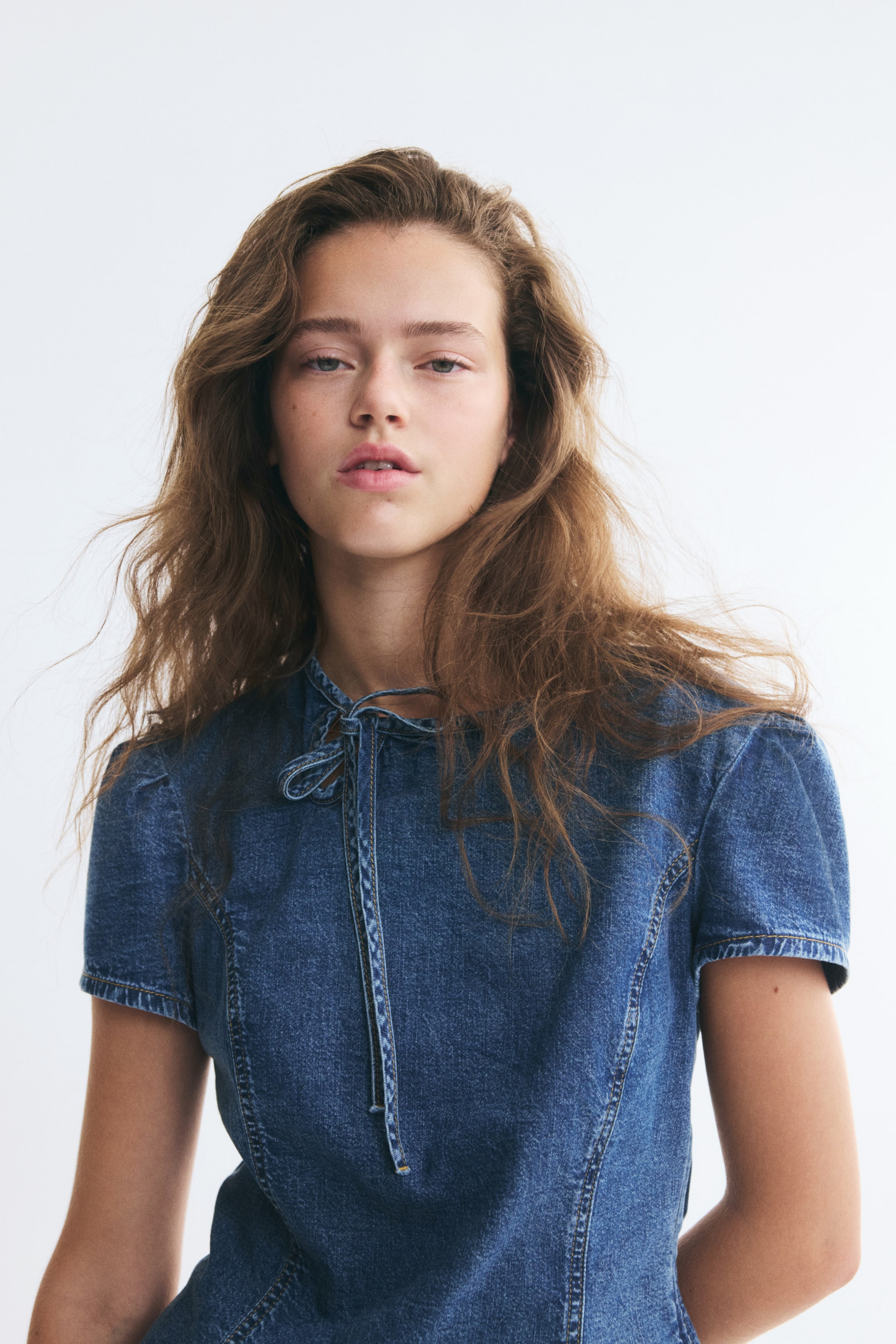 Xem ảnh lớn hơn: Người mẫu đang mặc một chiếc áo blouse denim ôm dáng, dáng ngắn. Áo có cổ tròn với chi tiết khoét giọt lệ và dây buộc mảnh ở một bên, tay áo phồng ngắn, cùng các đường may princess nổi chạy dọc phía trước.