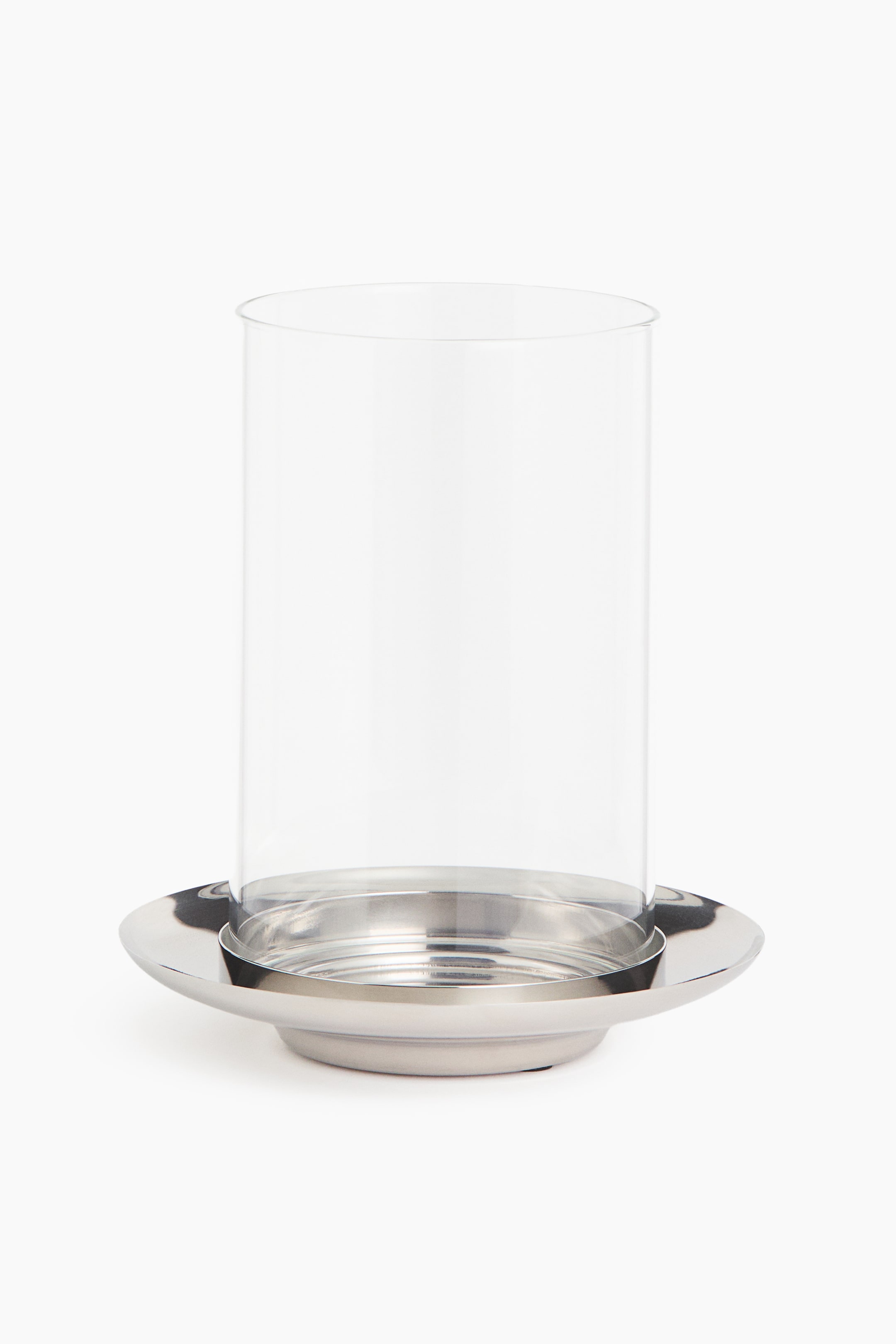 View larger image: A modern lantern is shown from a front angle, featuring a tall, cylindrical chimney in clear borosilicate glass resting on a polished stainless steel base. The base displays a flared, saucer-like rim with a recessed centre designed for holding a pillar candle.