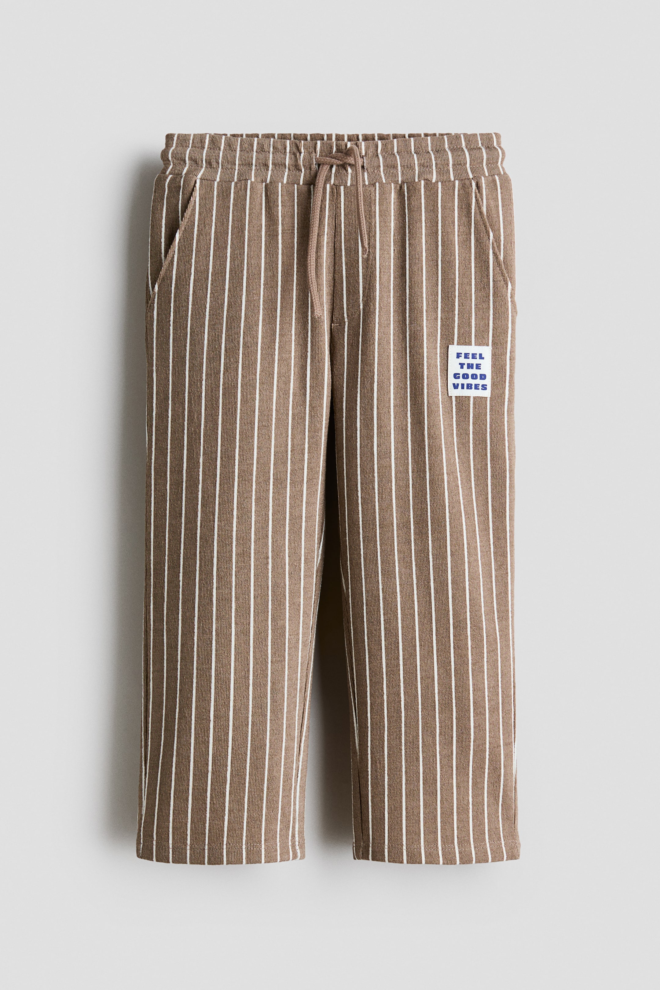 Visualizza l'immagine ingrandita: Pantaloni in jersey testurizzato, marroni con strisce verticali bianche, presentati frontalmente, con vita elasticizzata e coulisse, patta finta e tasche laterali diagonali. Una patch rettangolare con testo blu "FEEL THE GOOD VIBES" è applicata sulla parte superiore della gamba destra.