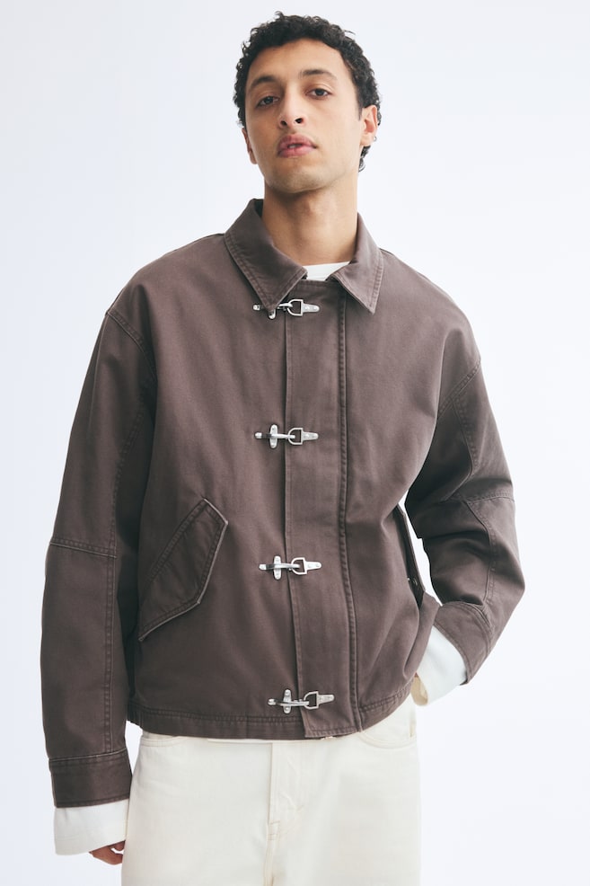 Die Model trÃ¤gt eine dunkelbraune Jacke aus Baumwoll-Canvas Ã¼ber einem weiÃen Oberteil und cremefarbenen Hosen. Die Jacke verfÃ¼gt Ã¼ber einen klassischen Kragen, Ã¼berschnittene Schultern, schrÃ¤ge aufgesetzte Pattentaschen auf der Vorderseite und einen Frontverschluss mit vier silberfarbenen MetallverschlÃ¼ssen. - 1