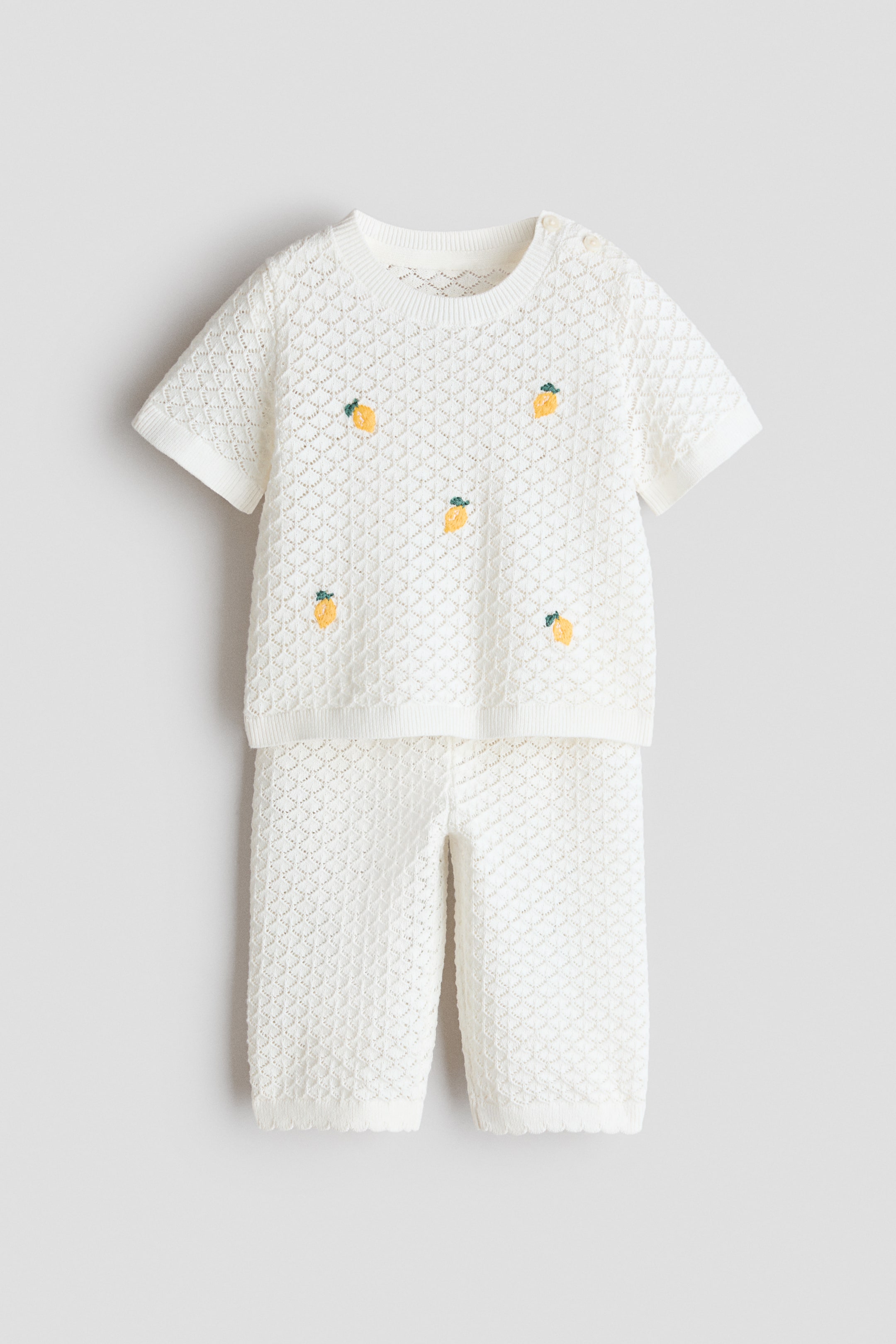 Ampliar la imagen: Un conjunto de algodón tejido en punto calado blanco presenta una blusa de manga corta y pantalones de 3/4 de largo, vistos desde el frente. La blusa cuenta con un escote redondo con ribete acanalado y botones en un lado, además de motivos bordados de limones amarillos, mientras que los pantalones muestran una cintura acanalada y aberturas de pierna con borde festoneado.