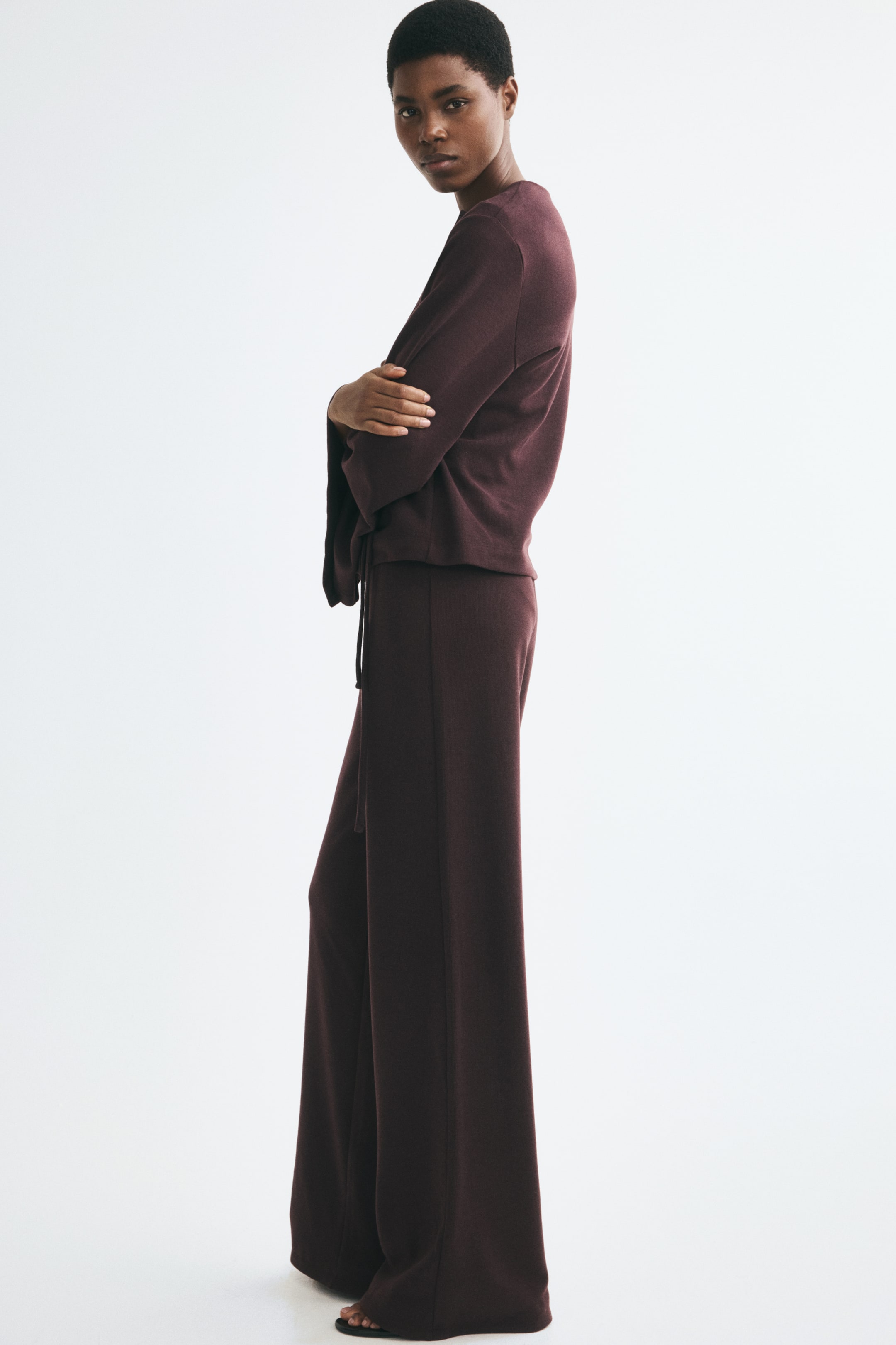 View larger image: The model is wearing relaxed-fit knit pants in a dark plum shade, paired with a matching long-sleeve top. The pants feature a soft texture, wide legs, and a drawstring waistband.