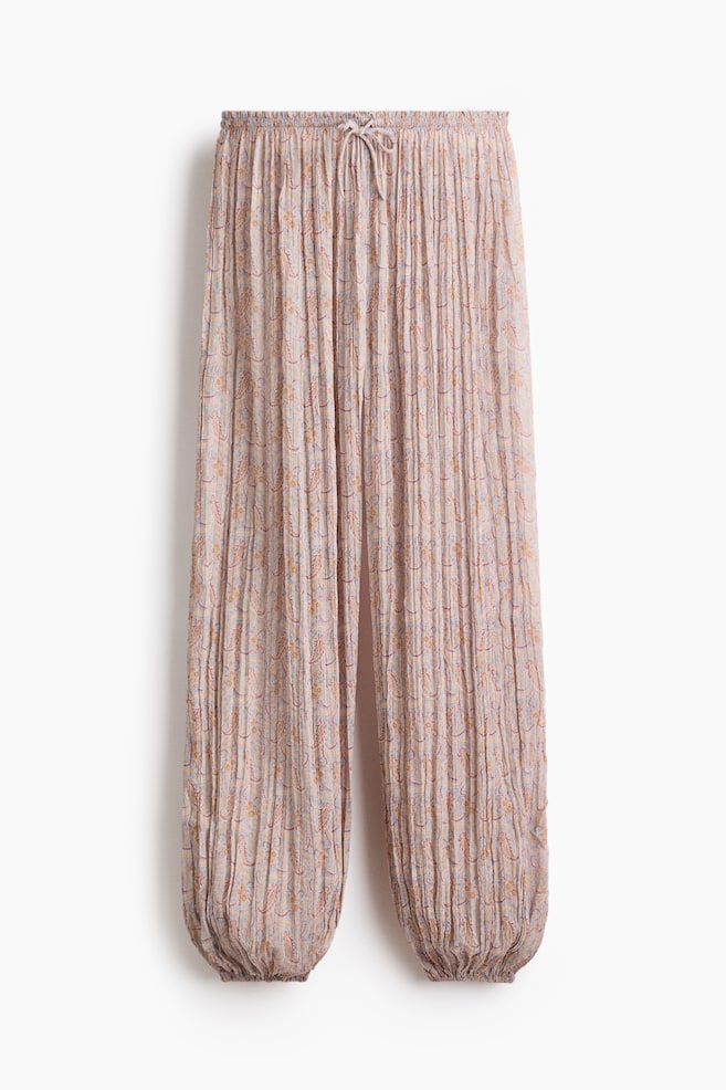 Los pantalones holgados de playa presentan un tejido transparente y arrugado con un sutil patrón ornamental, vistos desde el frente. Cuentan con una cintura elástica adornada con volantes y un cordón estrecho, terminado con pequeñas cuentas de plástico y lentejuelas perladas en los extremos, y piernas abombadas con elásticos estrechos en los dobladillos. - 1