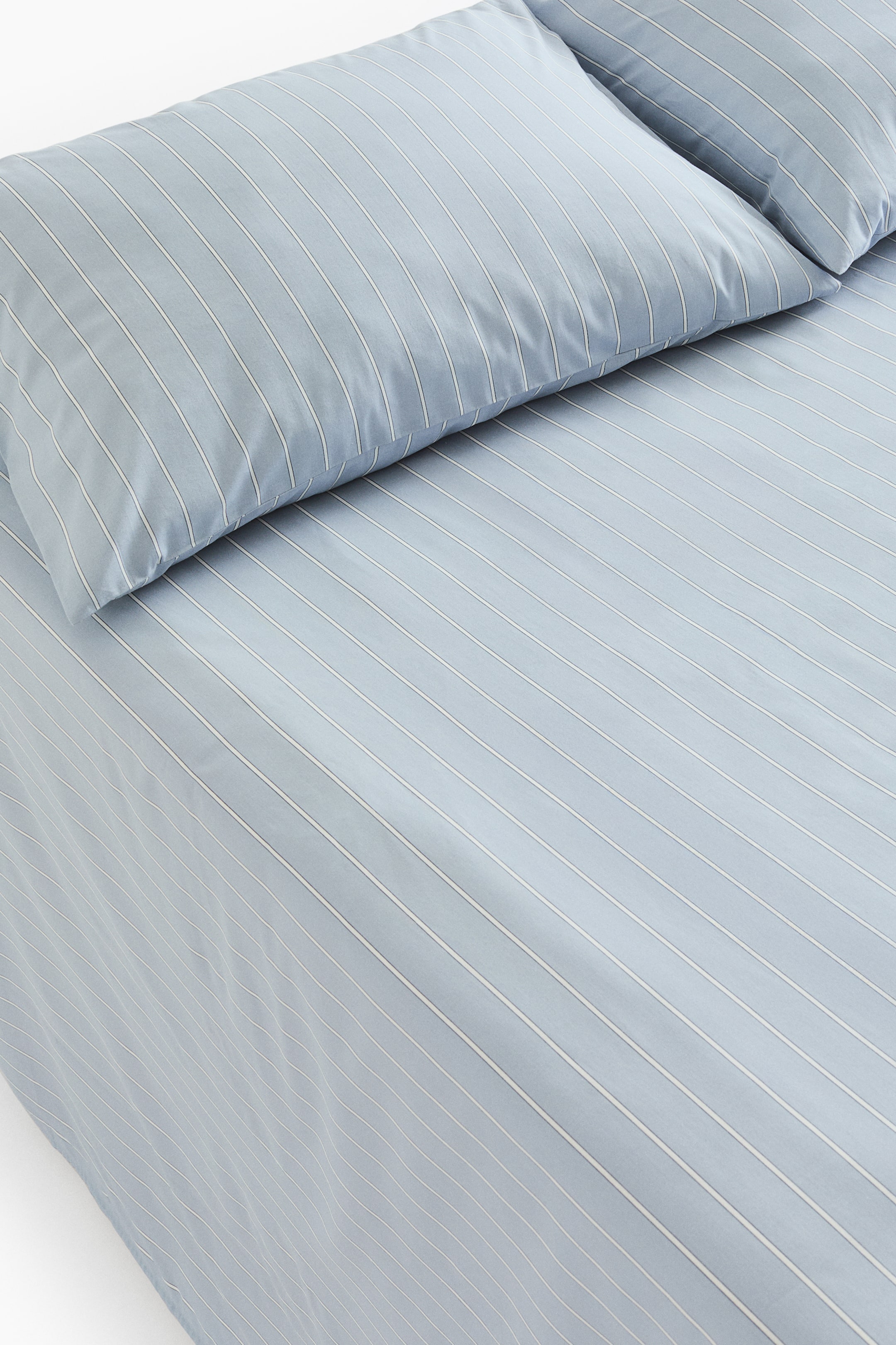 View larger image: A queen bed sheet set, made of a light blue soft cotton weave, is displaying a thin white striped print. One flat sheet is covering the surface with diagonal stripes, and two matching pillowcases are resting at an angle on top, showing vertical stripes.