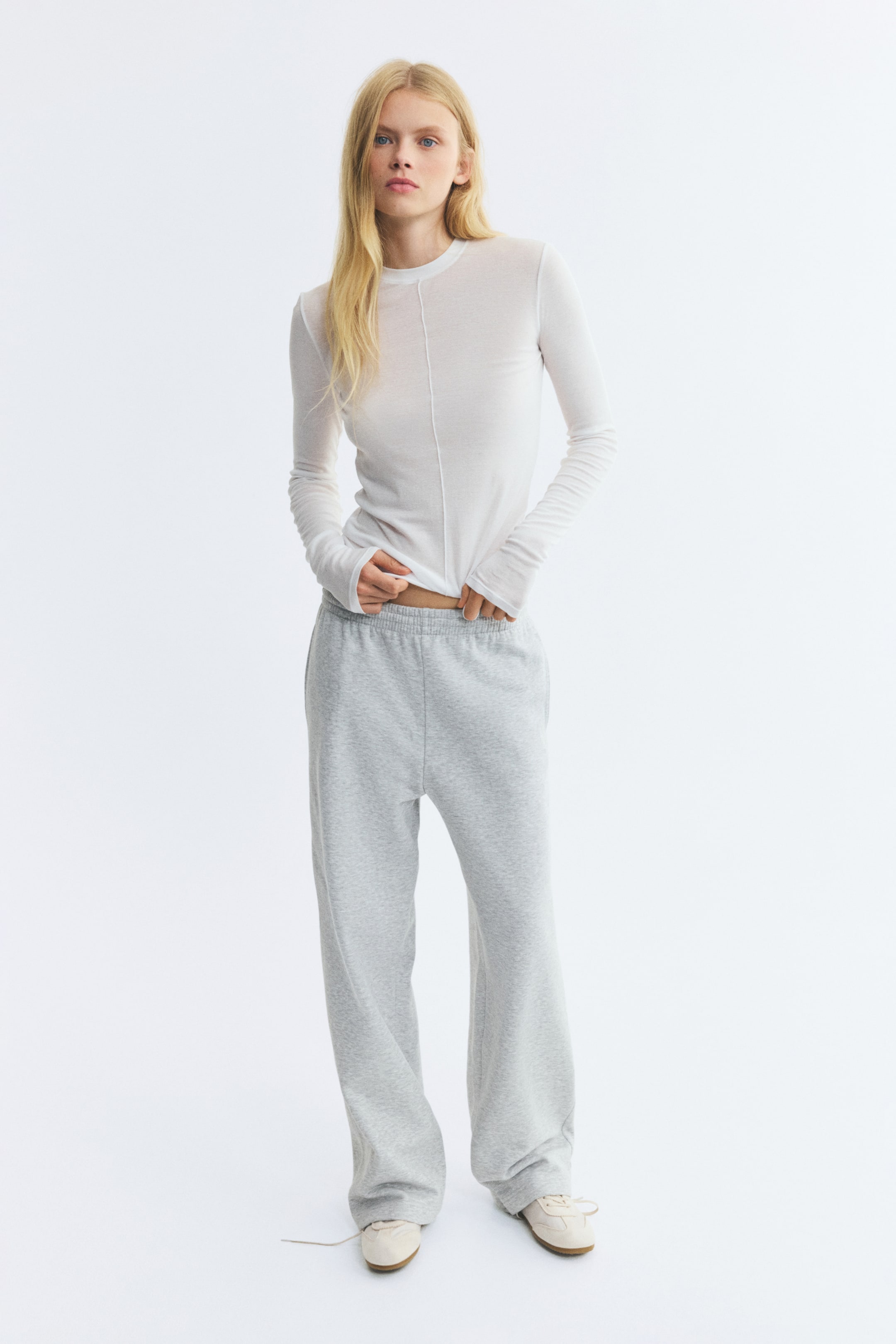 View larger image: Grey mélange track pants featuring an elasticated waistband and wide, straight legs are worn by the model. The model is standing and slightly lifting the hem of a semi-sheer white long-sleeved top to reveal the waistband of the pants.