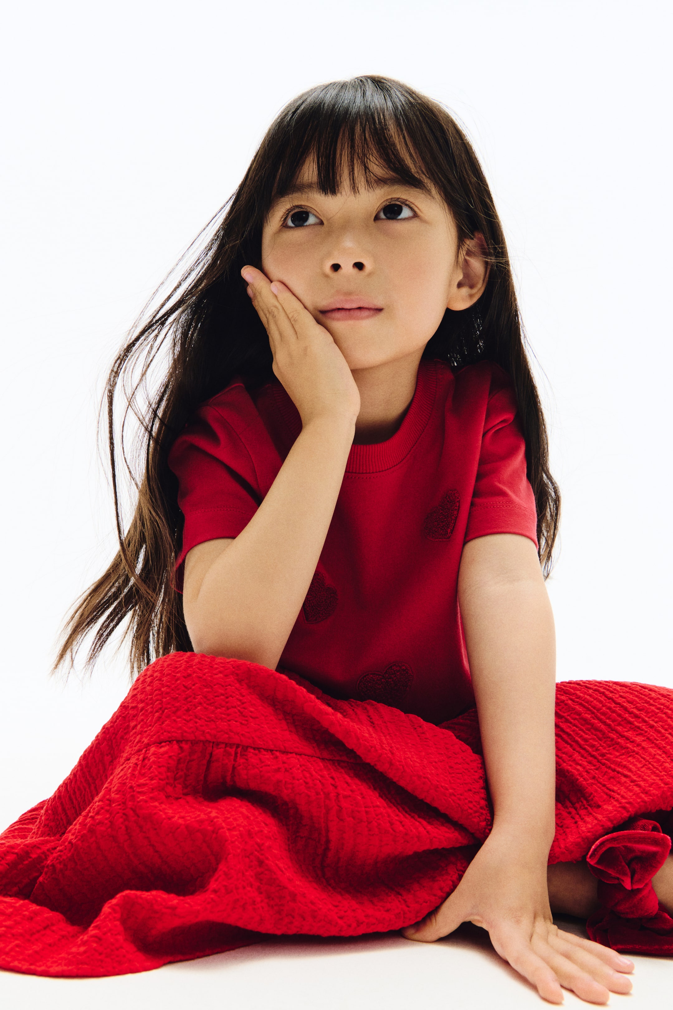 View larger image: A red outfit comprising a short-sleeved top and a textured skirt. The top features subtle, tone-on-tone heart patterns on the front, while the skirt is crafted from crinkled fabric that gently gathers around the wearer’s legs.