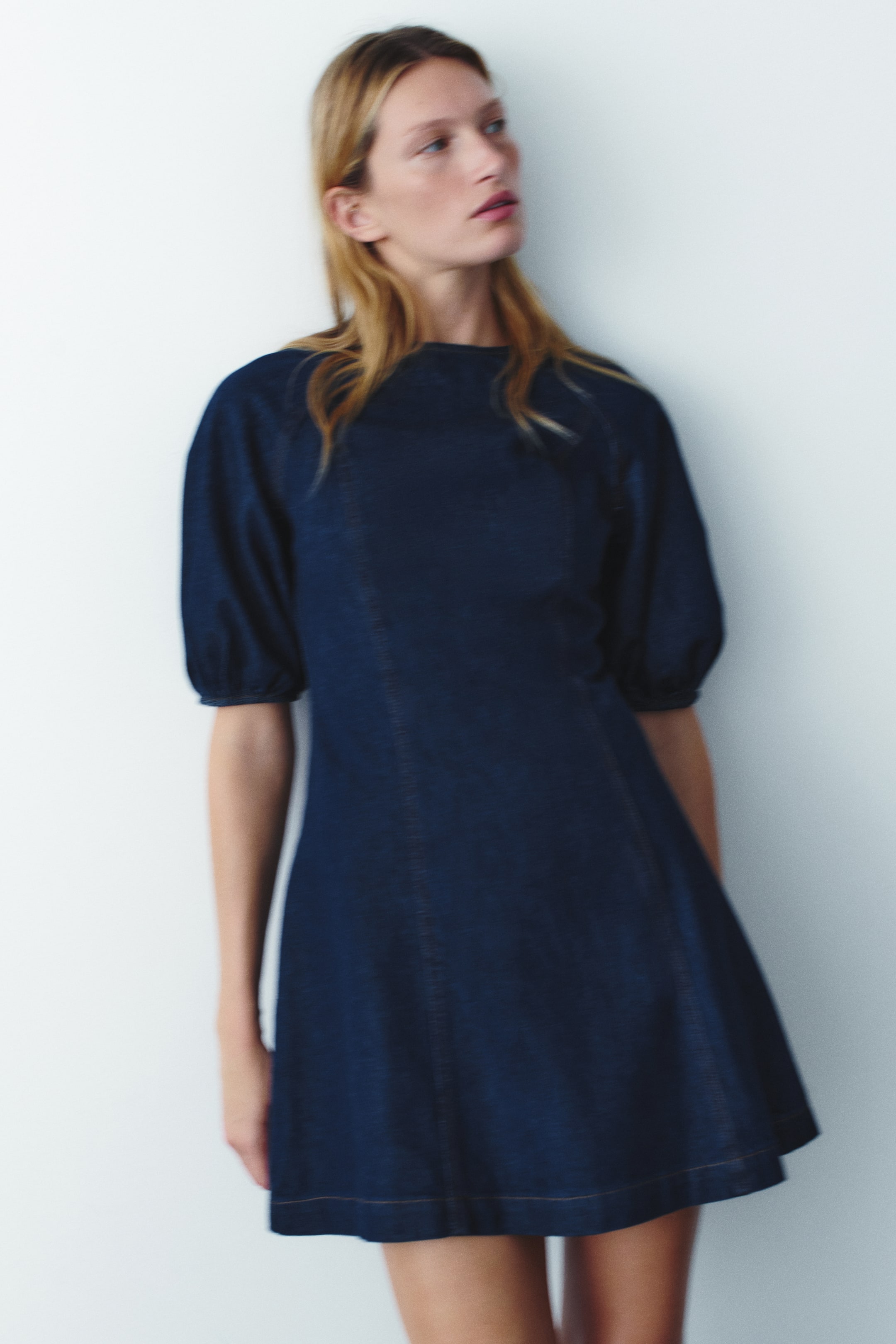 View larger image: The model is wearing a dark blue denim mini dress featuring a round neckline, short balloon sleeves with elasticized cuffs, a fitted bodice with princess seams, and a gently flared skirt.