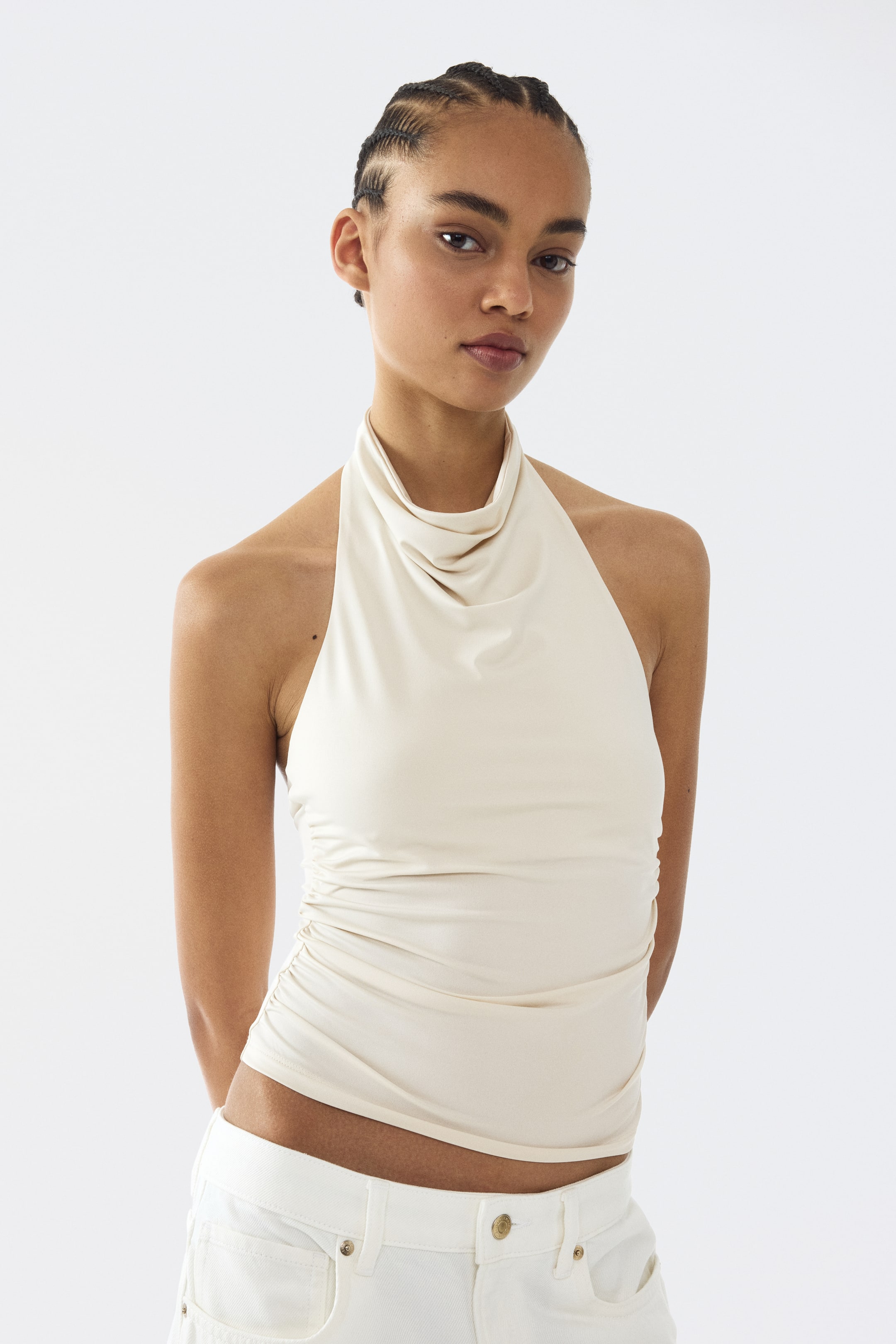 View larger image: A cream halter top showcasing a draped cowl neckline with ruched detailing along the left side. The top fits closely, offering a smooth texture.