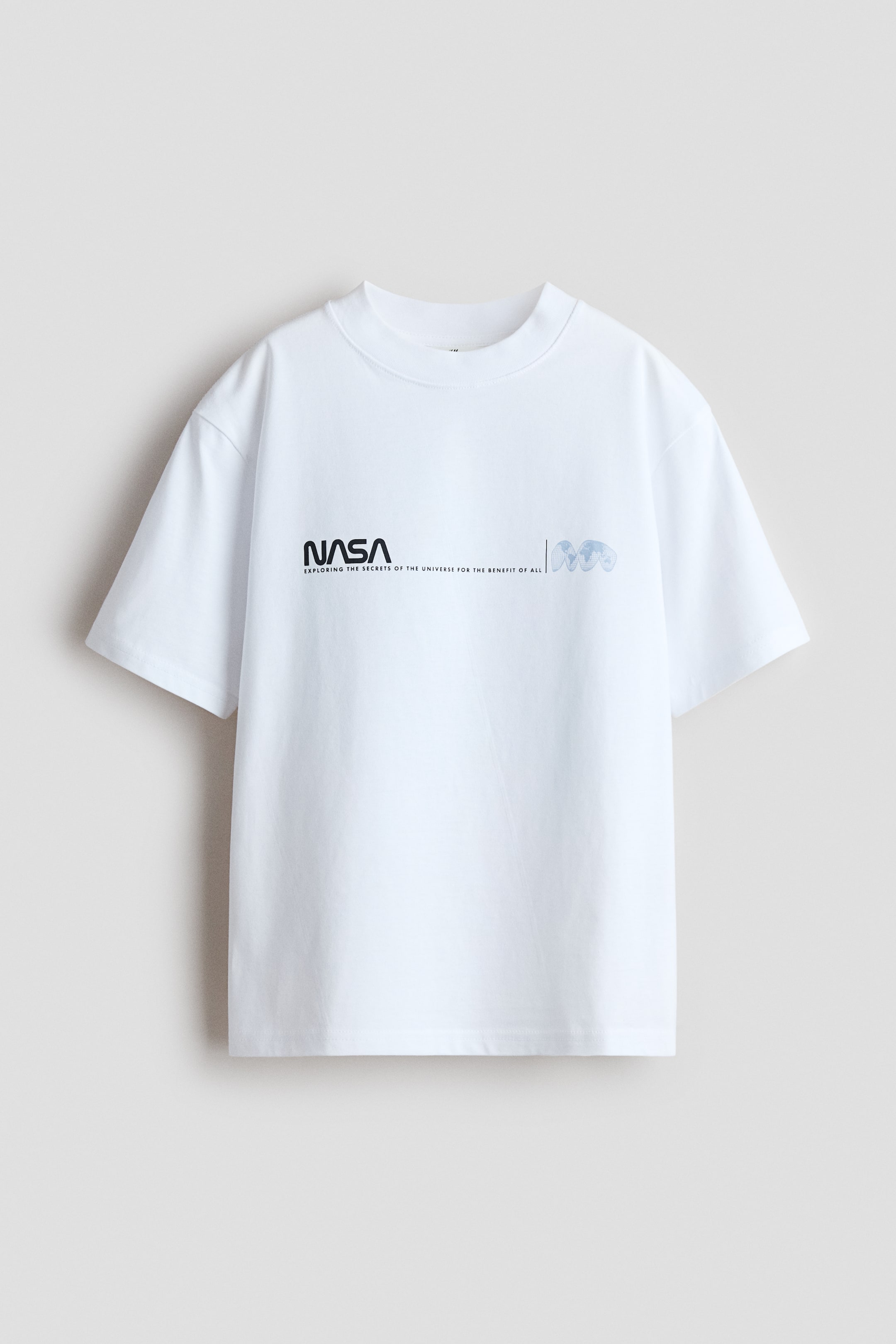 Visualizza l'immagine ingrandita: Una t-shirt in jersey bianco con stampa, a vestibilità ampia, è mostrata dal davanti, con un bordo a coste intorno al collo, spalle scese e una stampa "NASA" nera con testo aggiuntivo e tre forme astratte azzurro chiaro sul petto.