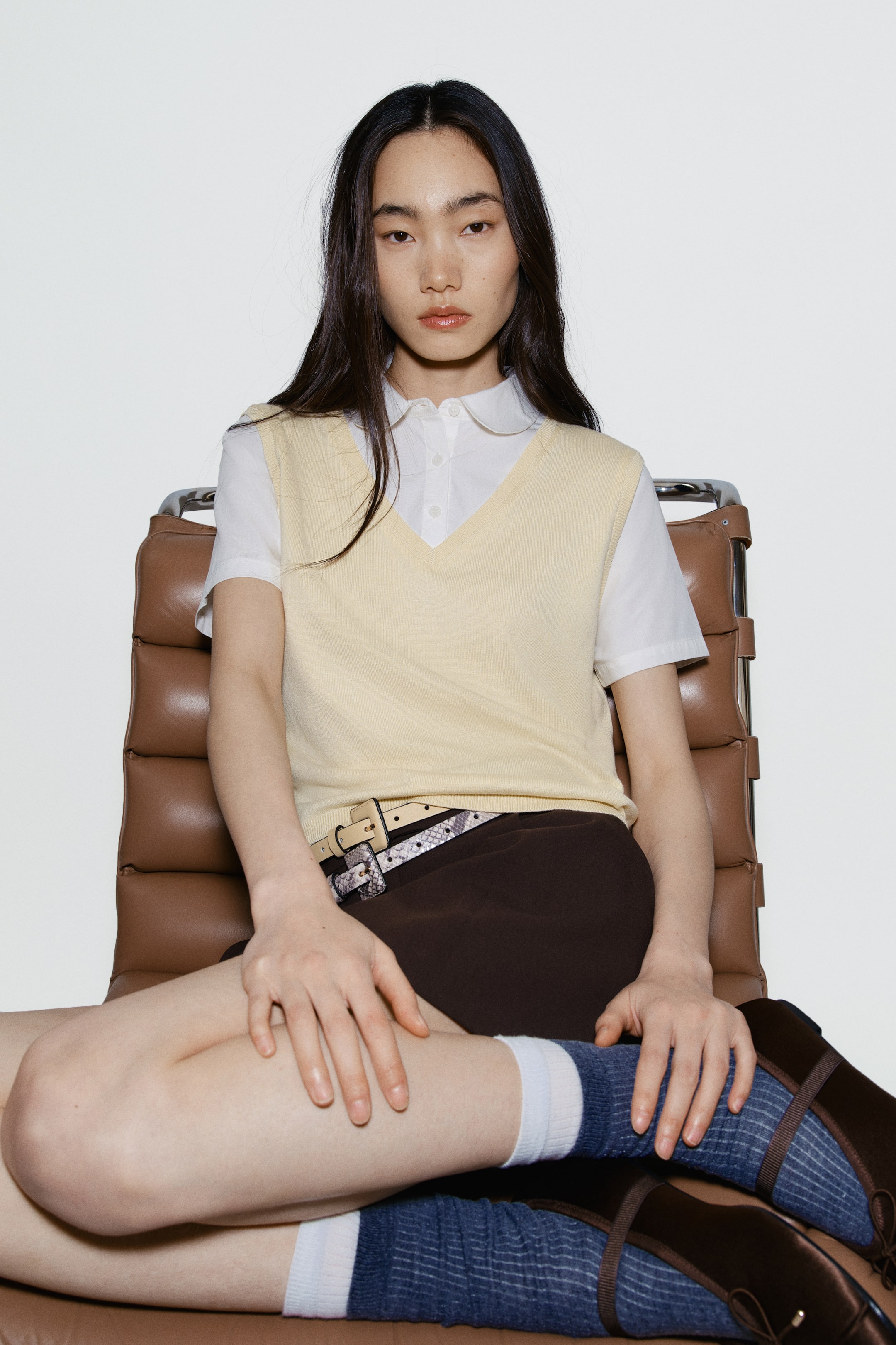 拡大画像を表示: ライトイエローのVネックセーターベストが、丸い襟の白い半袖シャツの上に重ねられています。ダークブラウンのショートパンツには、ライトカラーのスネークプリントベルト、青と白のストライプソックス、ダークブラウンのローファースタイルの靴が合わせられています。