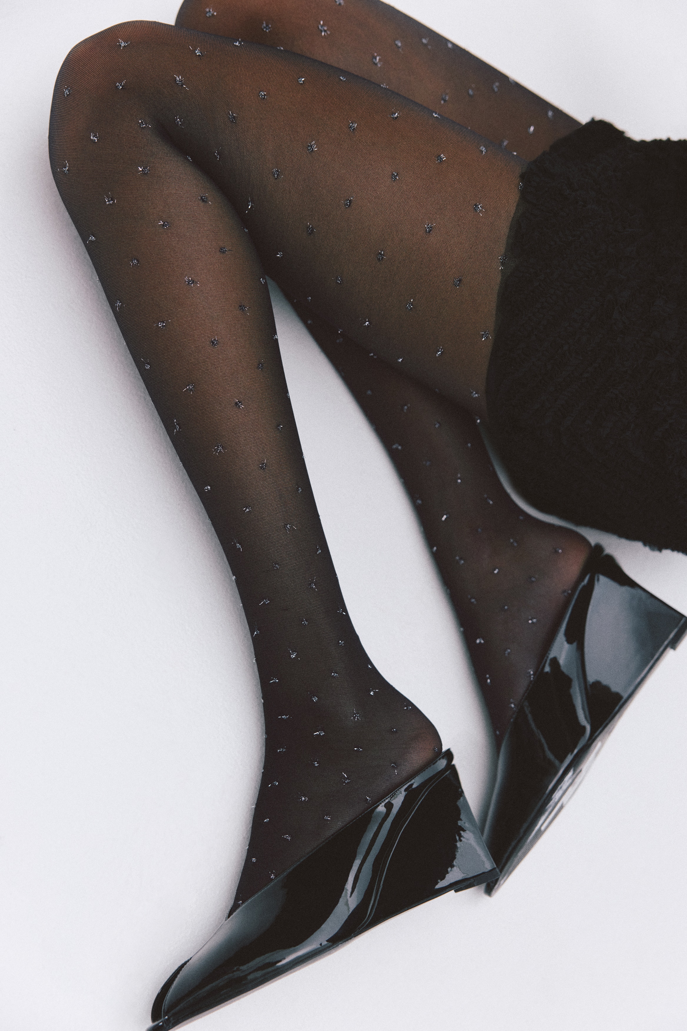 Un paio di collant neri è piegato e confezionato su un inserto di cartone bianco, mostrando piccoli frammenti metallici argentati sparsi sul tessuto trasparente. Una fascia di carta bianca avvolge il centro dei collant piegati, con il marchio "Tights" e "H&M".