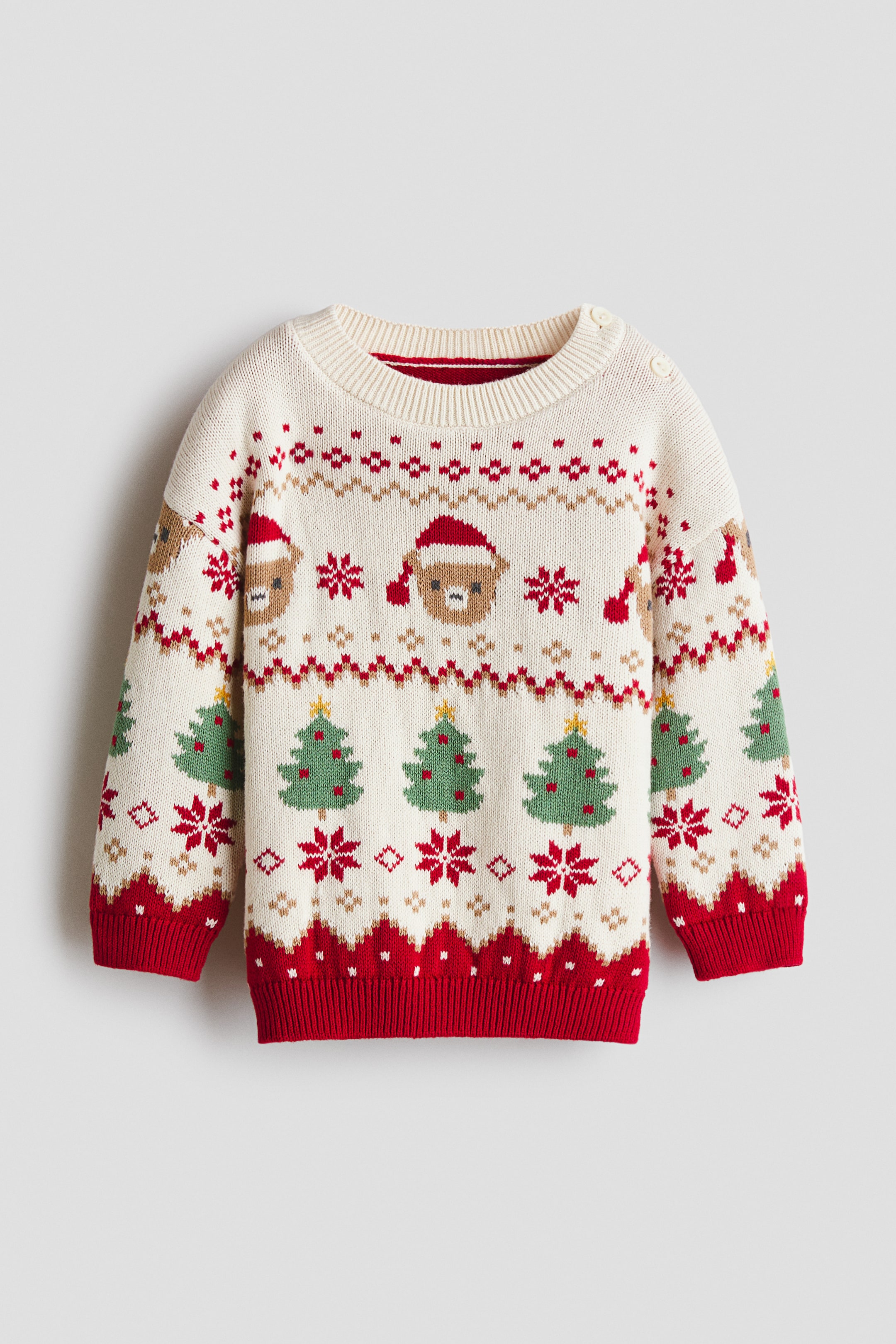 View larger image: A cream-coloured sweater is laid flat, displaying a festive Fair Isle pattern across the body. It features alternating brown bears in red Santa hats, green Christmas trees with yellow stars, and red snowflakes, and is finished with a ribbed crew neck, two white shoulder buttons, and red ribbed cuffs and hem.