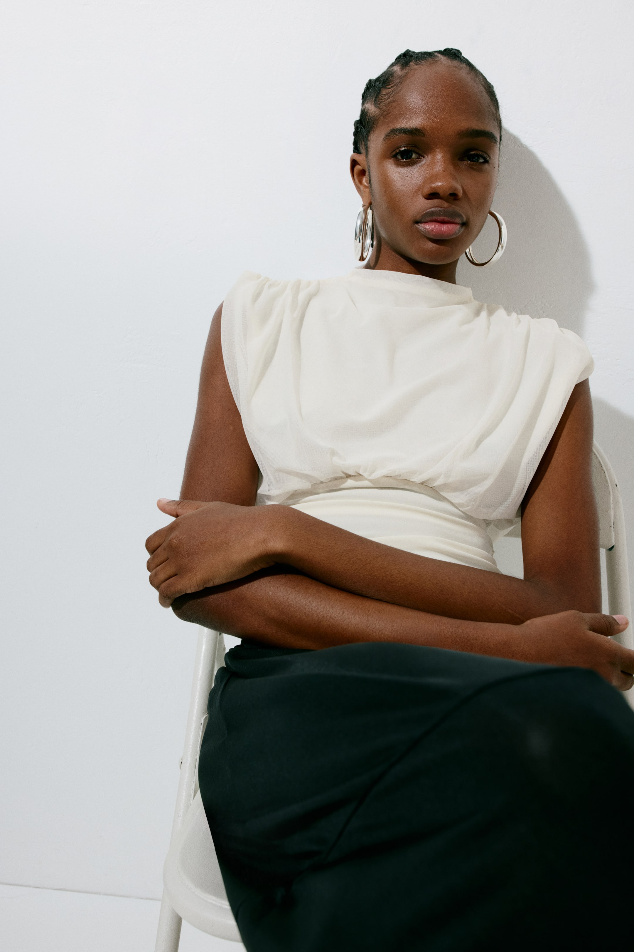 View larger image: A cream sleeveless top featuring a mock neck and gathered sheer fabric creating a structured shoulder. Its fitted bodice cinches at the waist.
