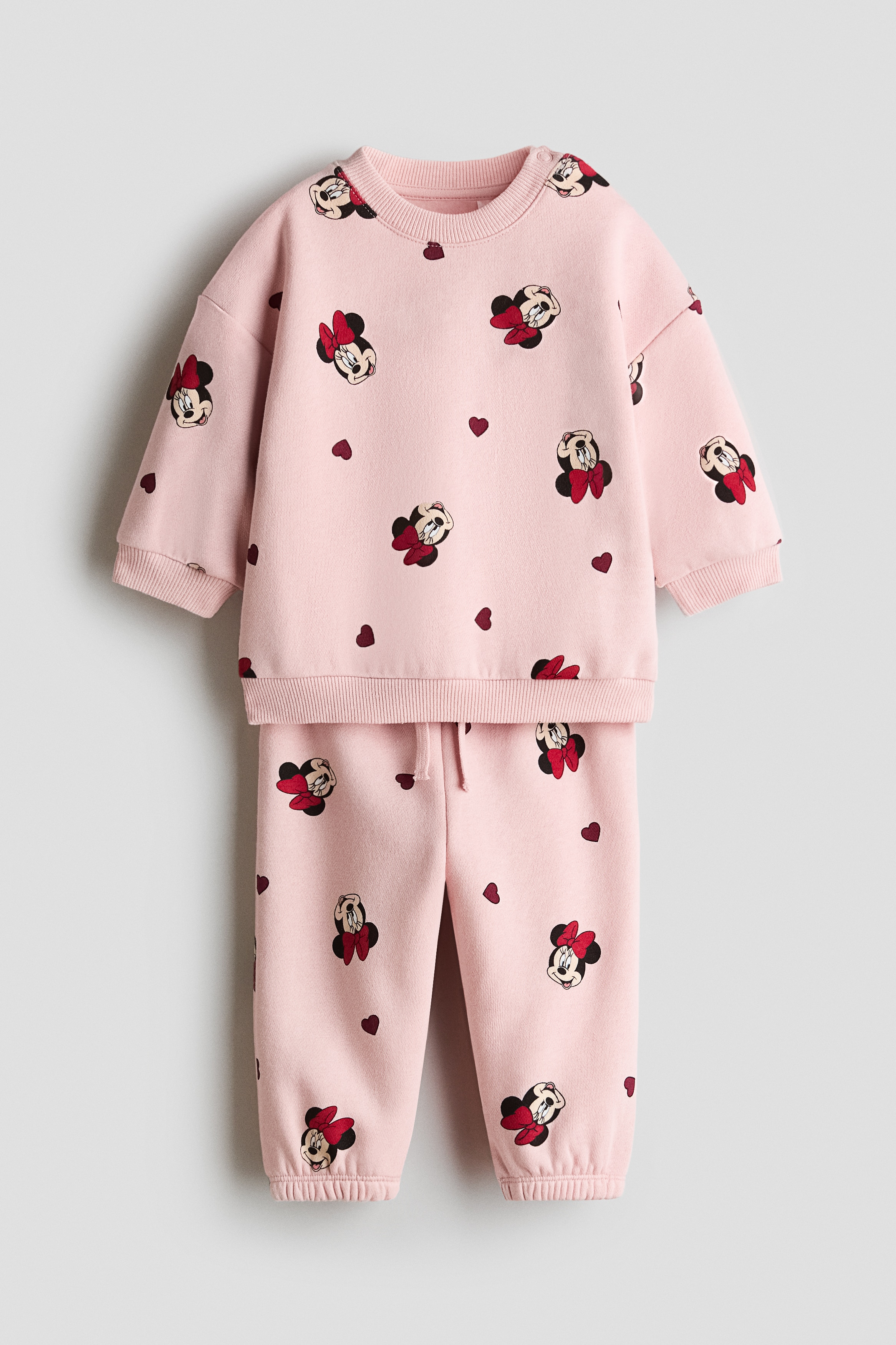 A 2-piece printed sweat fabric set in light pink is shown from the front, featuring an all-over pattern of Minnie Mouse faces with red bows and small red hearts. The set includes a long-sleeve sweatshirt with a ribbed crew neck and cuffs, paired with matching sweatpants featuring gathered ankles.