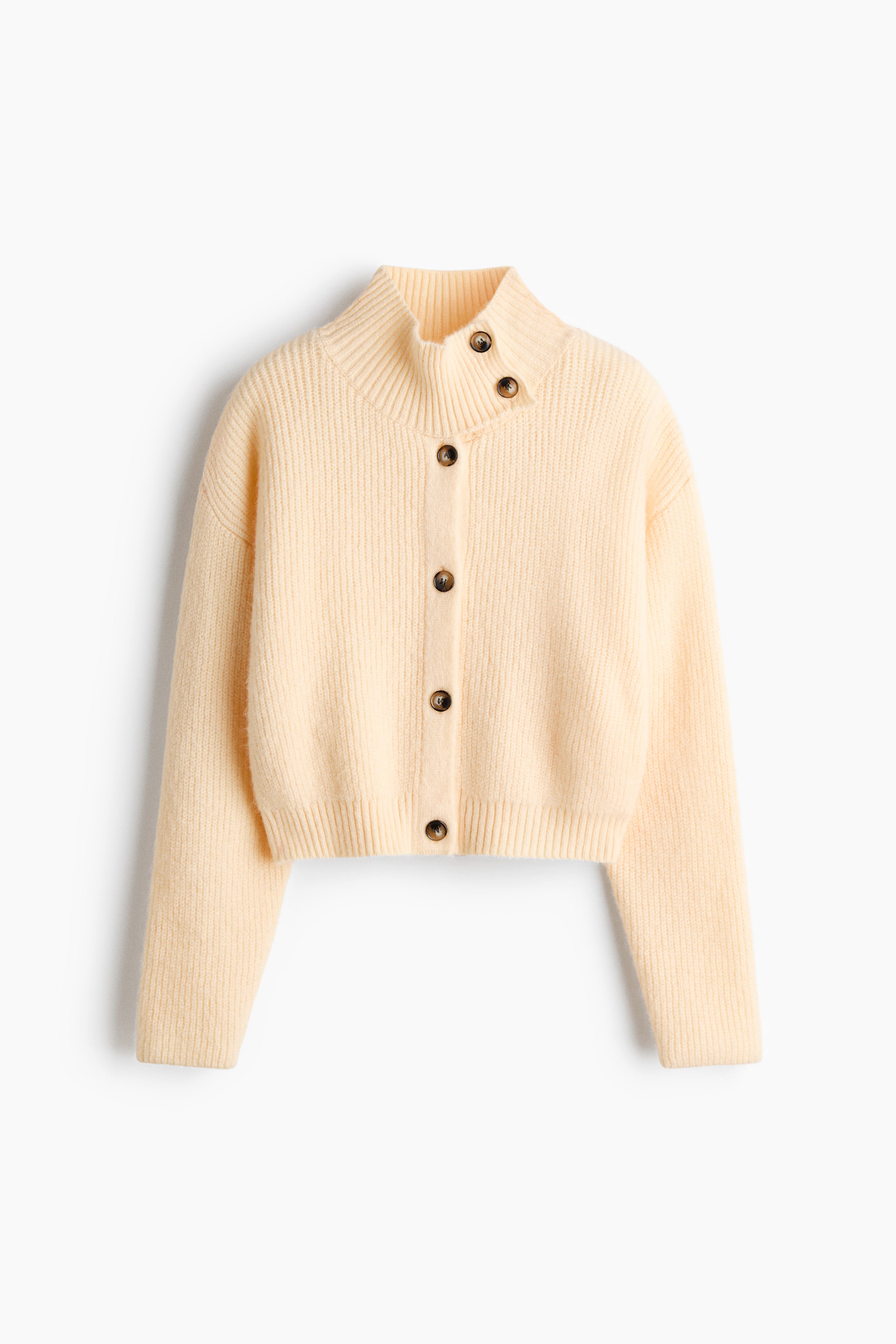Größeres Bild ansehen: Eine verkürzte, hellcremefarbene Strickjacke mit Rippenstruktur ist von vorne zu sehen. Sie verfügt über einen hohen Stehkragen mit asymmetrischem, geknöpftem Kragen und überschnittenen Schultern. Dunkelbraune Schildpattknöpfe verlaufen mittig entlang der Knopfleiste und runden das Design mit Knopfleiste ab.
