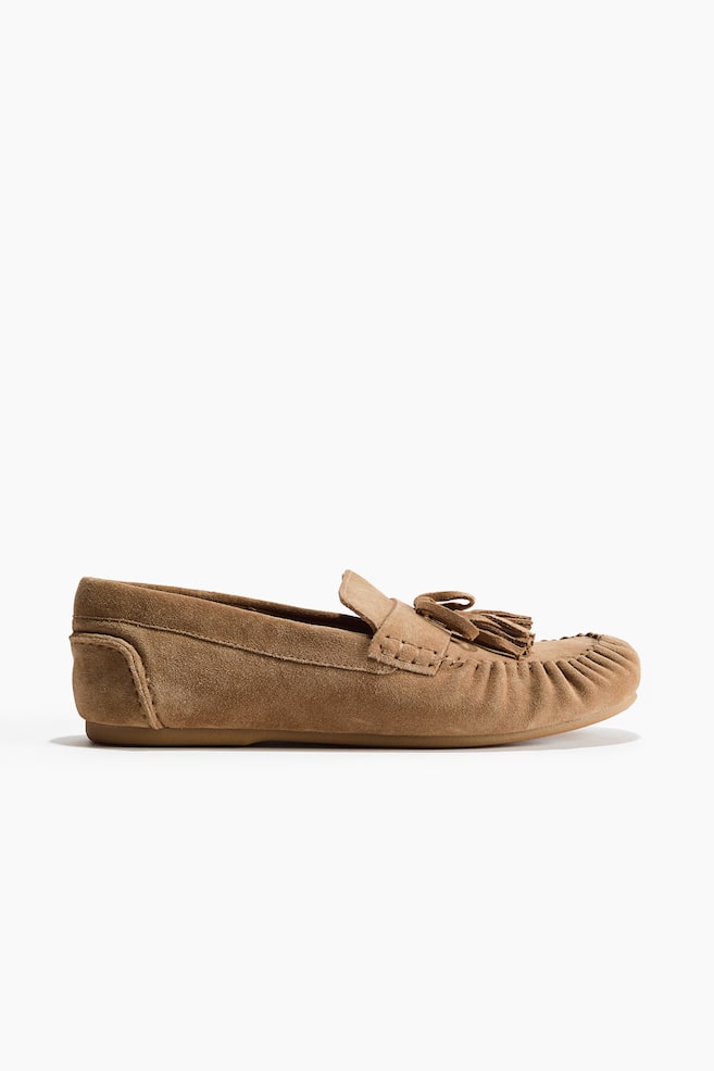 A single tan suede moccasin loafer is presenting its side profile, featuring a rounded, gathered moccasin toe, a vamp strap, and a decorative tassel detail. - 1