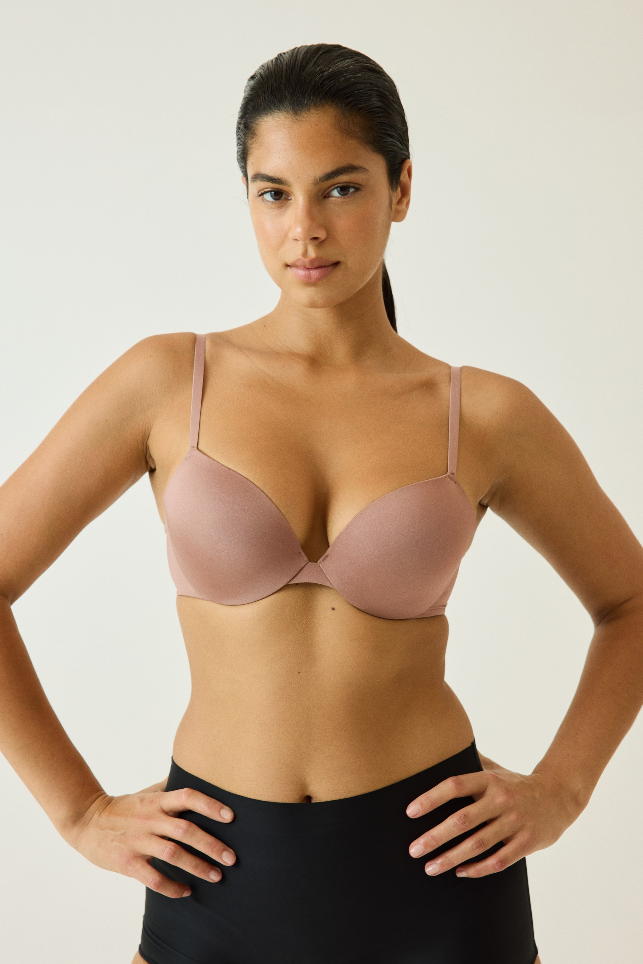 View larger image: A dusty pink microfibre push-up bra with seamless, padded cups and thin shoulder straps is worn by the model. The model is posing with hands on hips, wearing the bra paired with high-waisted black briefs.