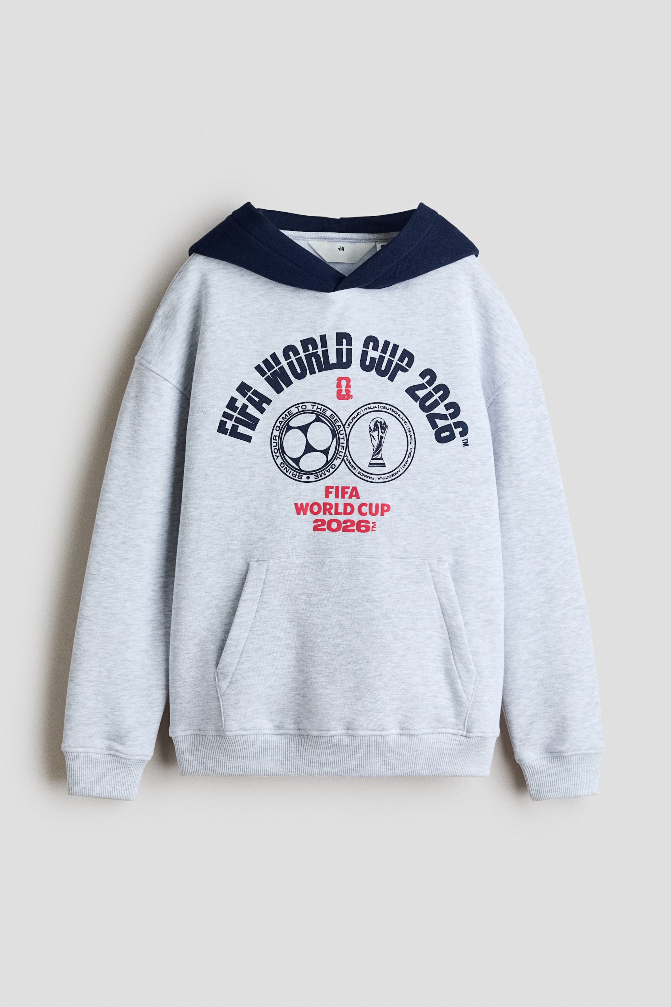 Agrandir l'image: Un sweat à capuche gris clair chiné est présenté de face, avec une capuche doublée de bleu marine contrastant. Un grand imprimé graphique est centré sur la poitrine, affichant "FIFA WORLD CUP 2026" en bleu marine et rouge, accompagné de deux emblèmes circulaires représentant un ballon de football et un trophée. Une poche kangourou est visible à la taille avant, et les manches longues se terminent par des poignets côtelés, avec un ourlet côtelé à la taille.