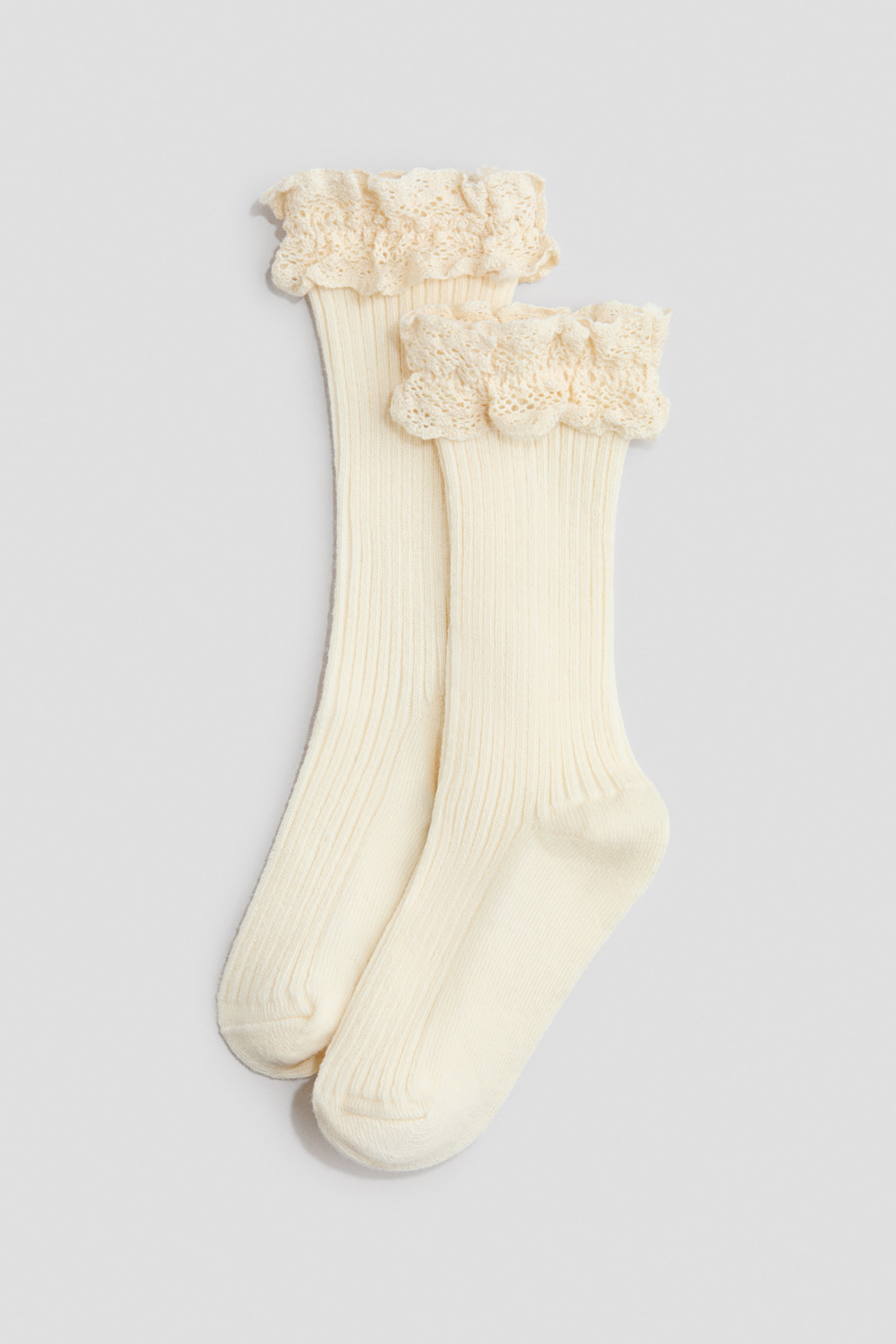 Ver imagem maior: Um par de meias de cor creme apresenta a sua parte frontal, com uma textura de malha fina canelada ao longo da perna. Frufrus delicados de renda de croché adornam os punhos superiores.