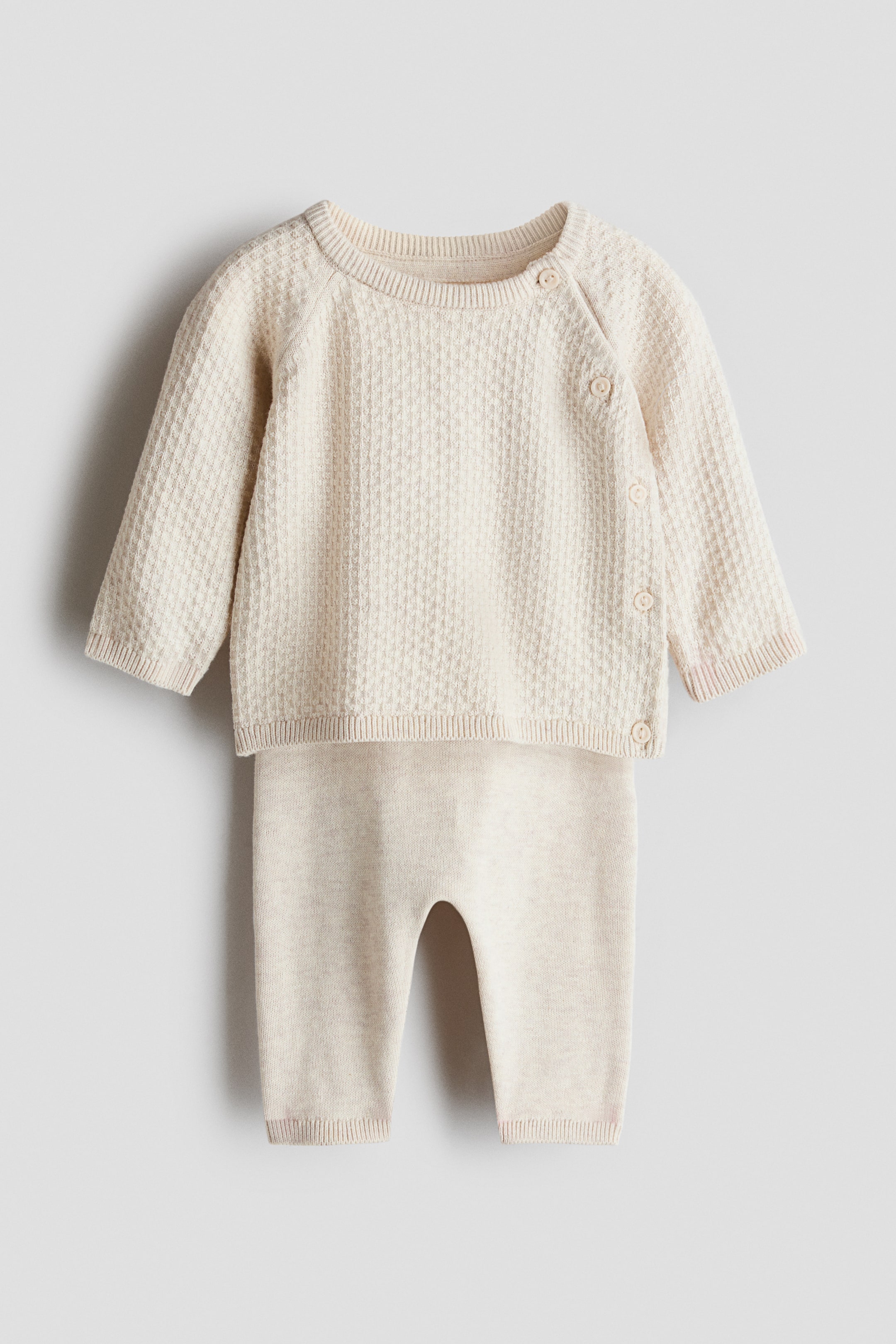 Ver imagen más grande: Un conjunto de dos piezas en color beige claro muestra un jersey de punto texturizado y unos pantalones de punto suave, ambos de frente. El jersey presenta un cuello redondo acanalado, mangas raglán largas con puños acanalados y cierre de botones a lo largo de un hombro y por el lateral. Los pantalones muestran una cintura elástica acanalada y bajos acanalados.