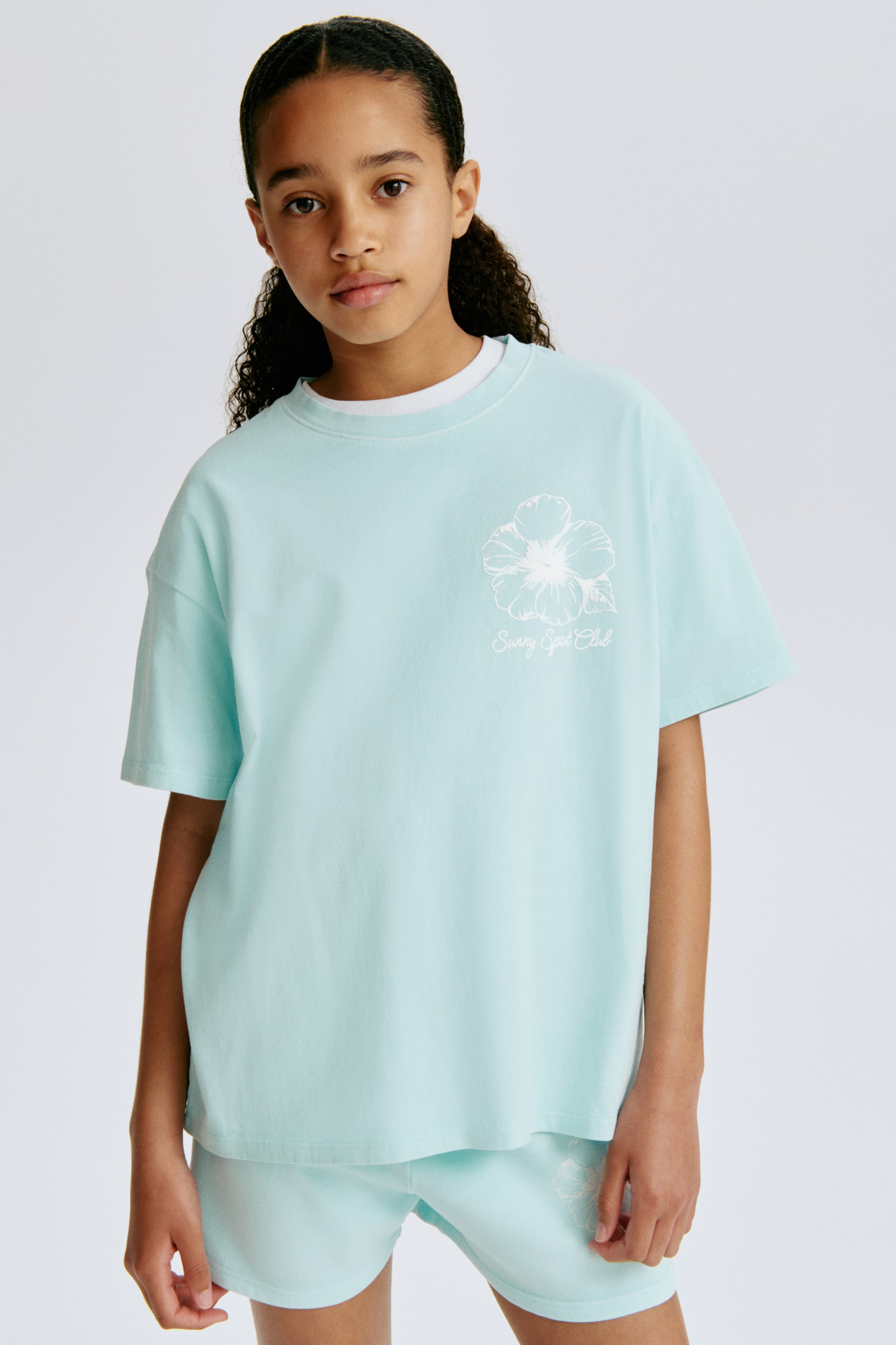 Grotere afbeelding bekijken: Een lichtblauw T-shirt met korte mouwen heeft een witte bloemenprint met de tekst "Sunny Spot Club" op de linkerborst, met een witte laag zichtbaar bij de ronde hals. Bijpassende lichtblauwe shorts zijn ook zichtbaar, met een subtiele bloemenprint op het linkerbeen.