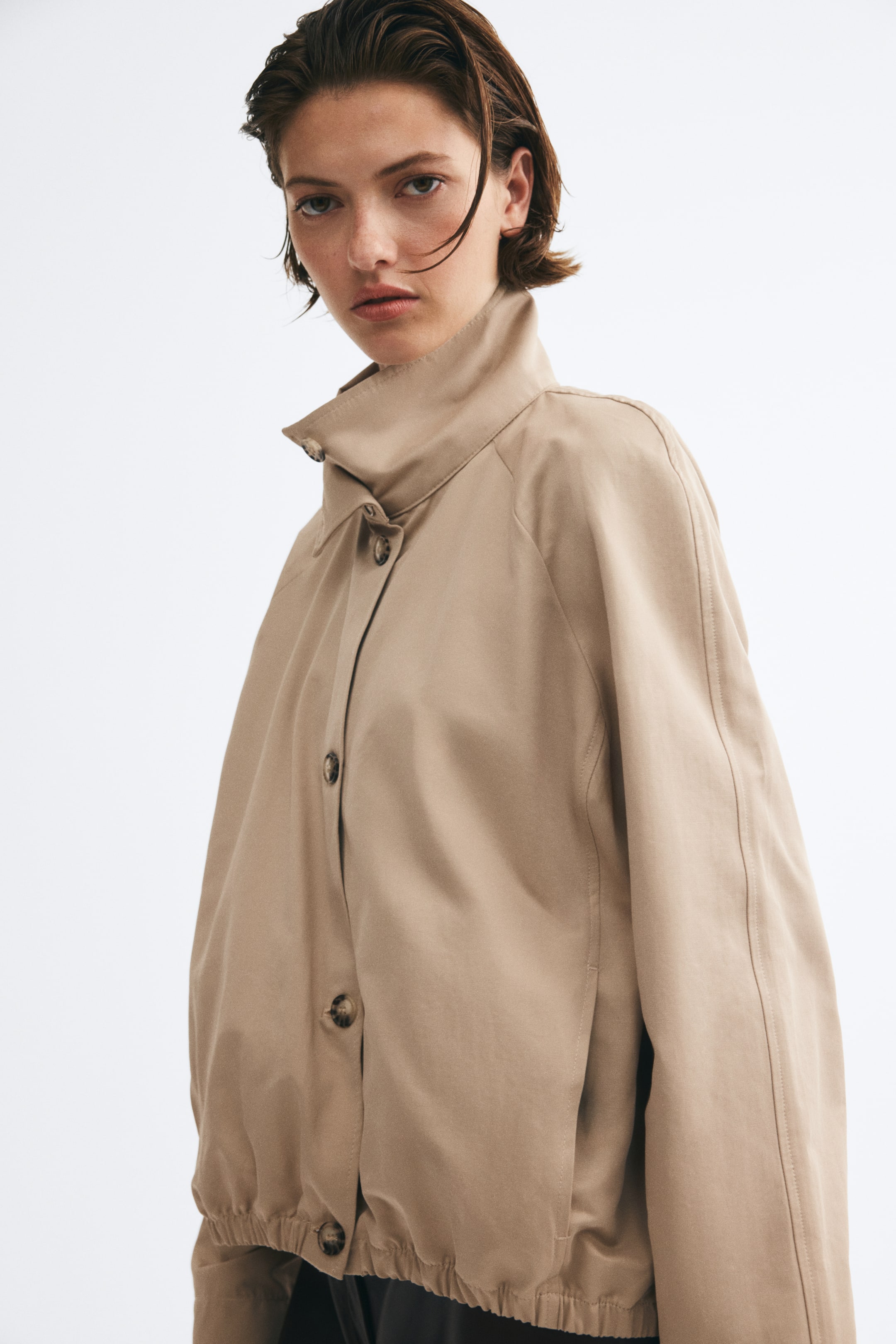 Visa större bild: En oversized beige jacka i vävt tyg bärs av modellen, med hög polokrage, knäppning framtill och raglanärmar. Plagget är stylat med en elastisk bubble-ärm nederkant som rynkas vid midjan.