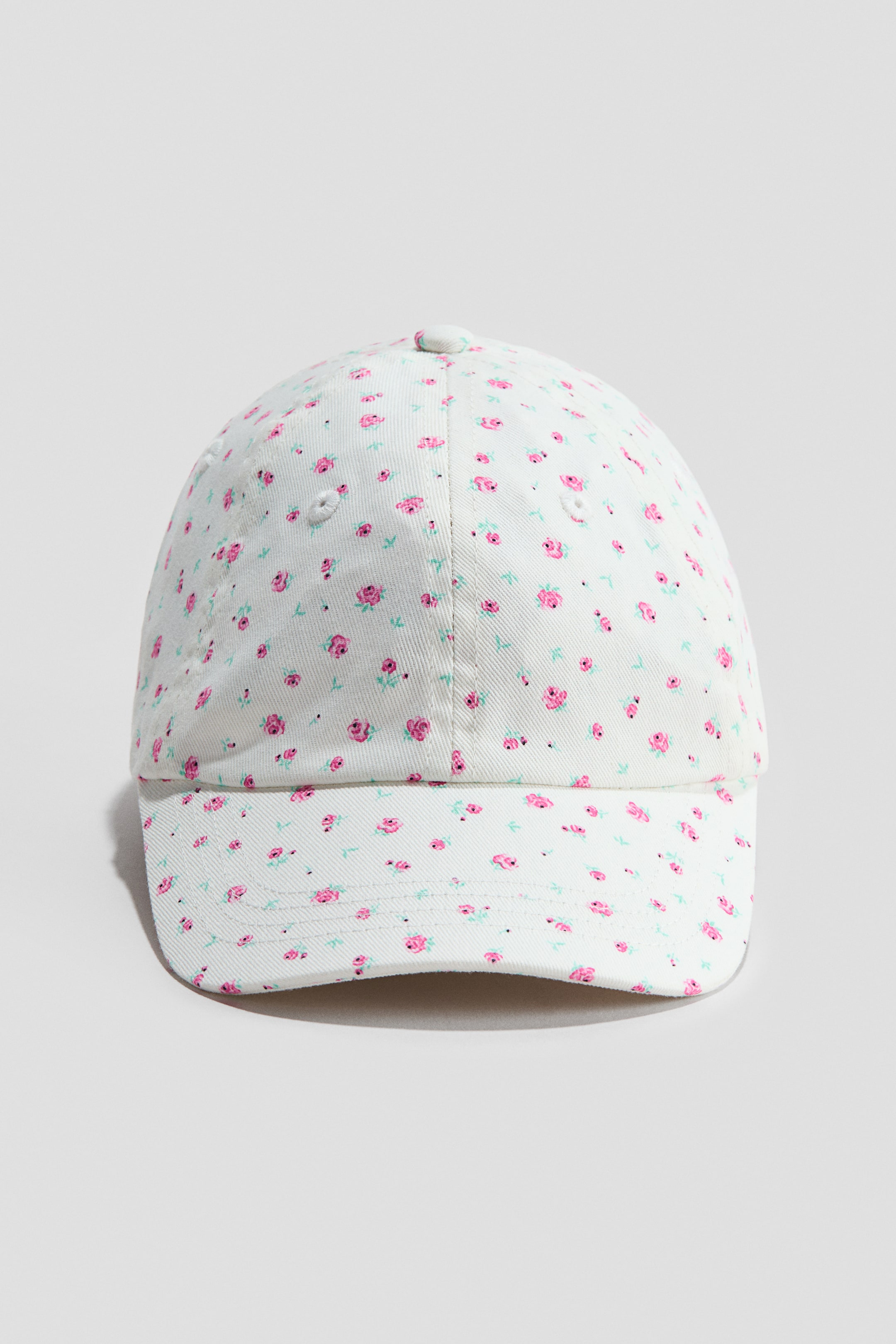 View larger image: The cap is facing forward, crafted from cotton twill, and features a repeating floral pattern of small pink roses and teal leaves set against a white background. Embroidered eyelets are visible on the top panels.