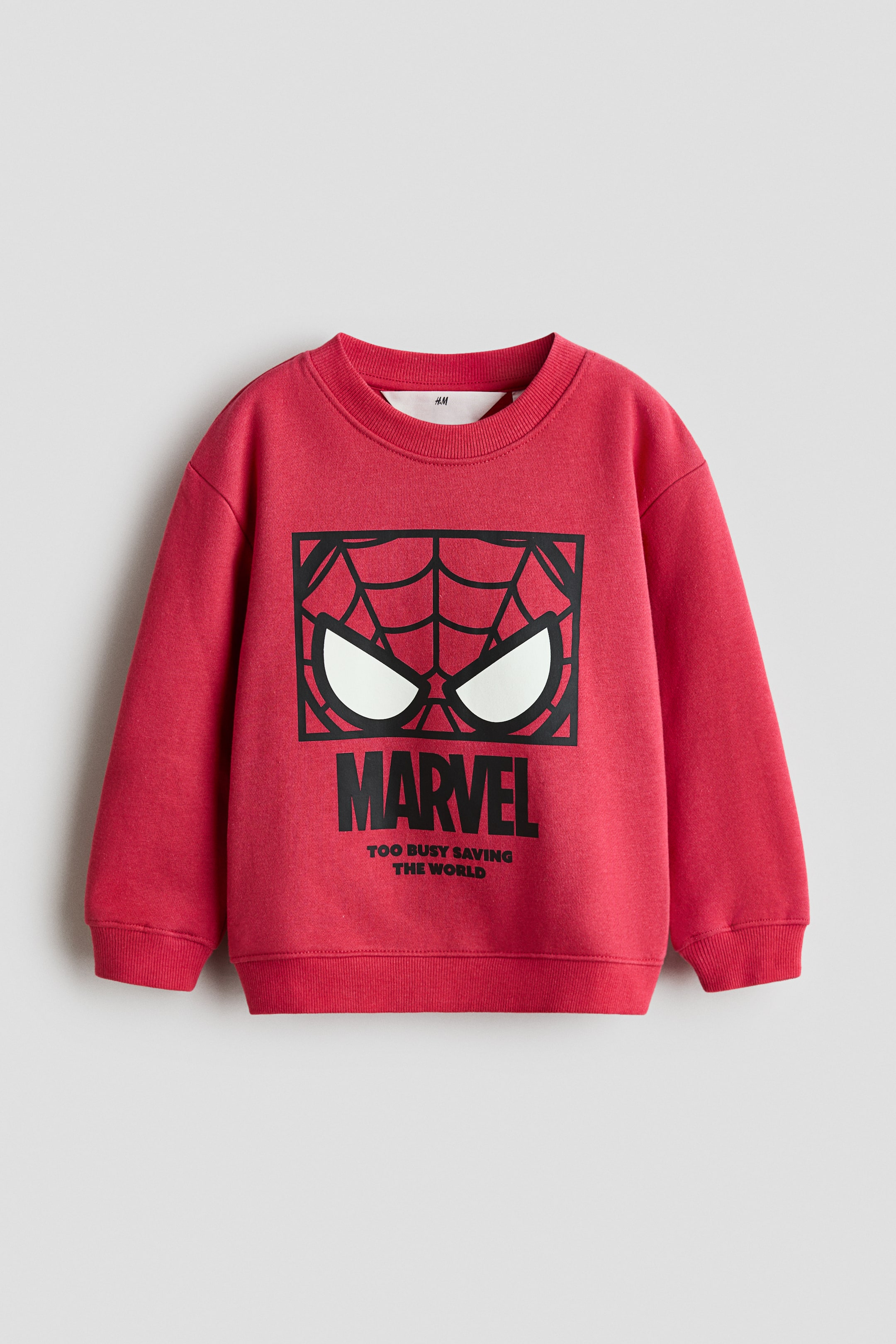 View larger image: A red sweatshirt is shown facing front, featuring a ribbed crew neck, long sleeves with ribbed cuffs, and a ribbed hem. Prominently displayed on the front is a large black graphic depicting a spider web pattern with two white eye shapes, the word 'MARVEL' in bold black letters, and 'TOO BUSY SAVING THE WORLD' in smaller black text below it.