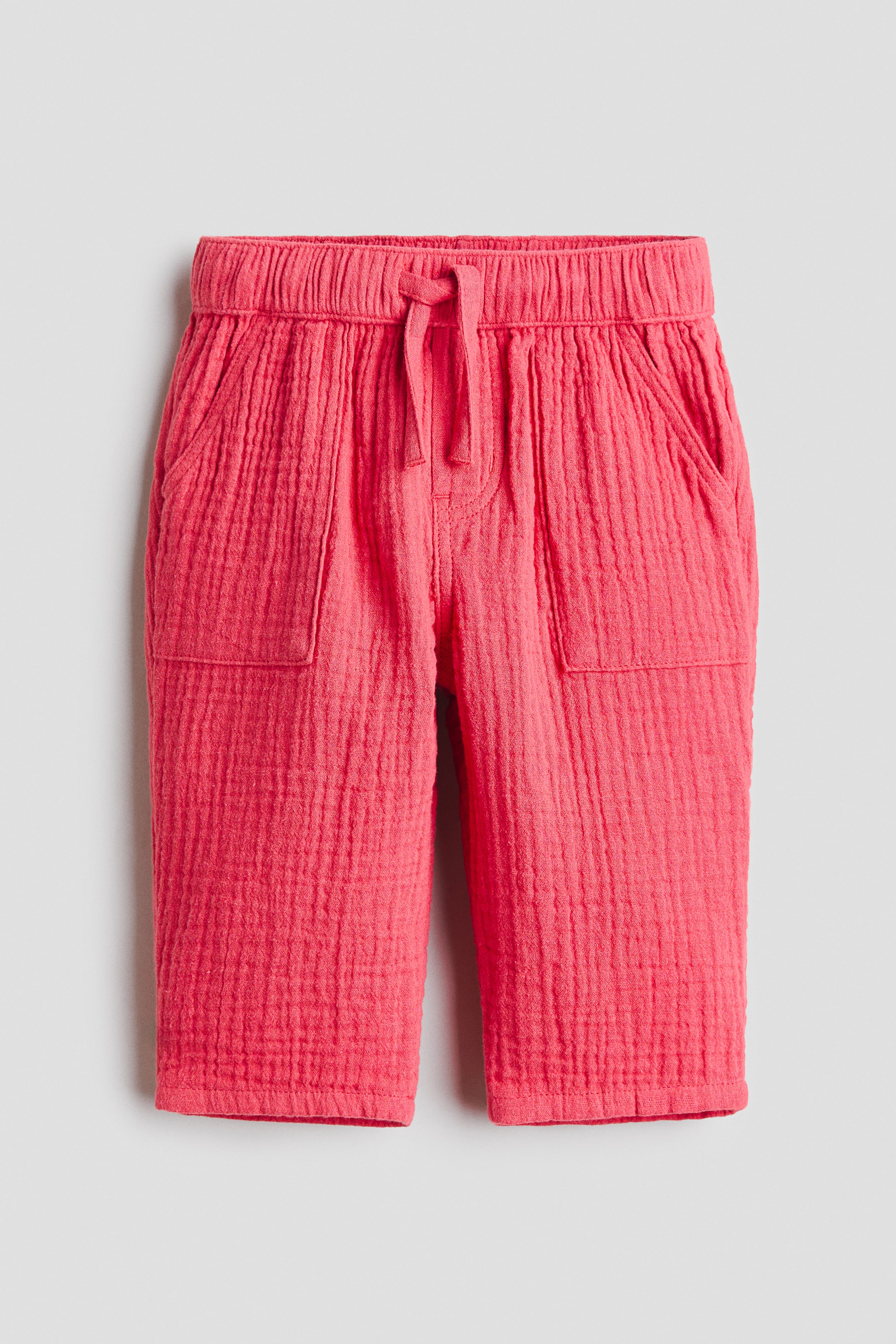 View larger image: Coral pink trousers crafted from soft, crinkled cotton muslin are showcased from the front. They feature an elasticated drawstring waist tied in a bow, a faux fly, two patch front pockets, and tapered legs.