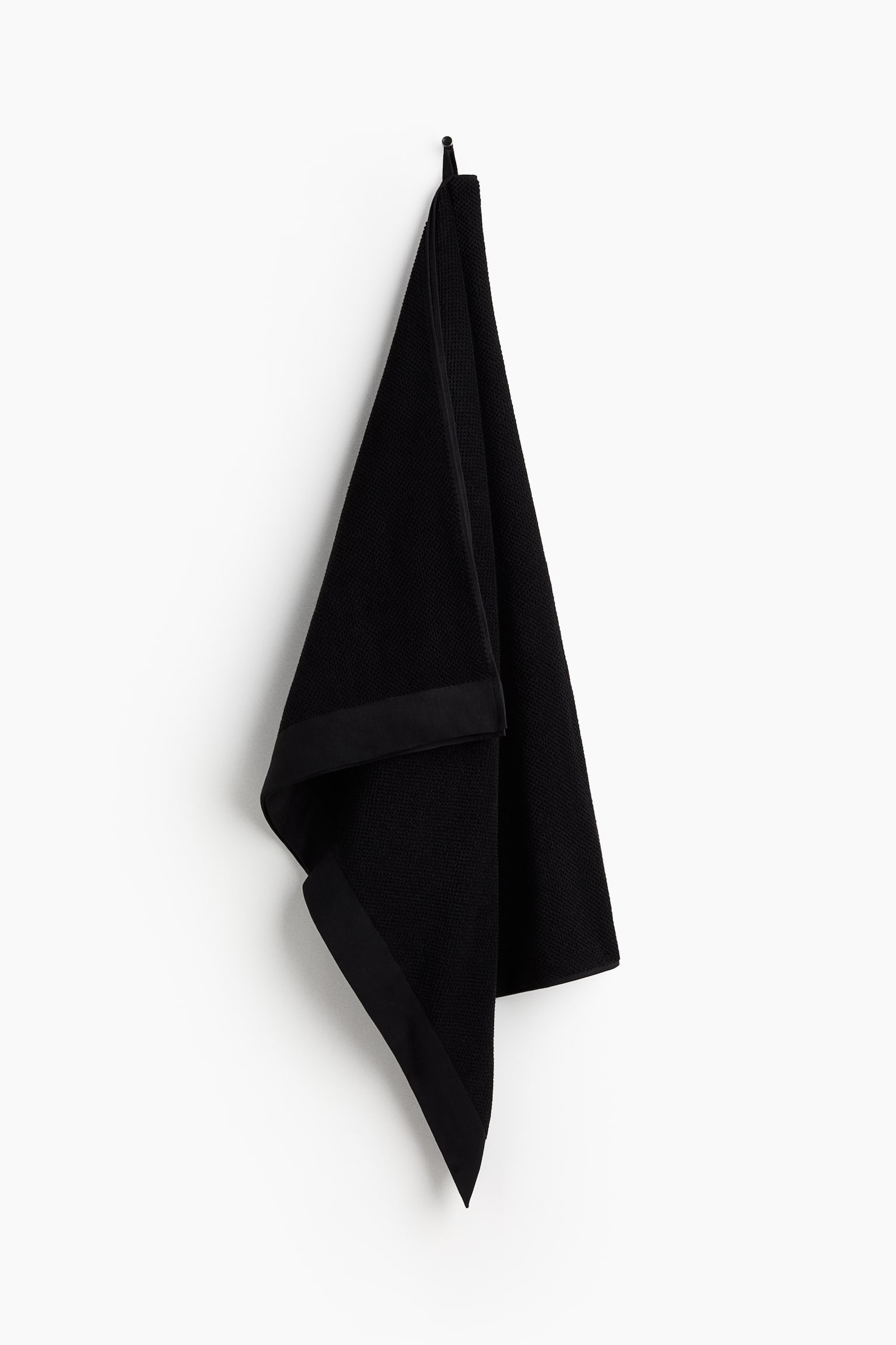 A black bath sheet is hanging folded from a loop at its top edge. It is showcasing a textured cotton terry material and a smooth border along its folded edge. - 1