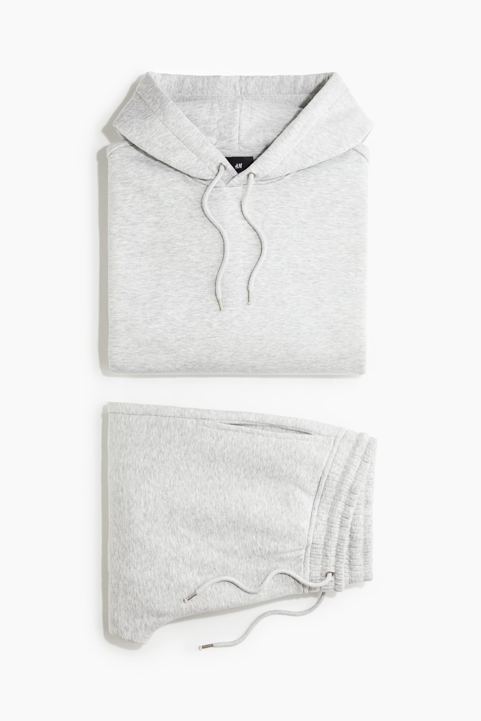 A light grey melange sweat set is laid folded flat from a front-facing angle, showcasing a hoodie on top and matching track pants below. The hoodie features a lined hood with hanging drawstrings, and the folded track pants display an elasticated waistband with a matching drawstring.