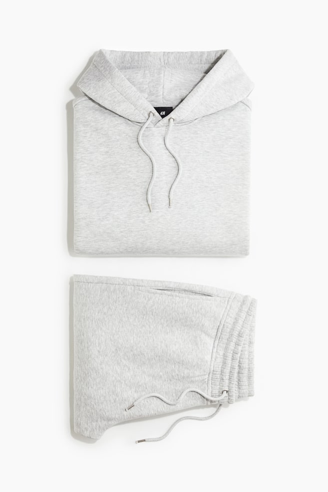 A light grey melange sweat set is laid folded flat from a front-facing angle, showcasing a hoodie on top and matching track pants below. The hoodie features a lined hood with hanging drawstrings, and the folded track pants display an elasticated waistband with a matching drawstring. - 1