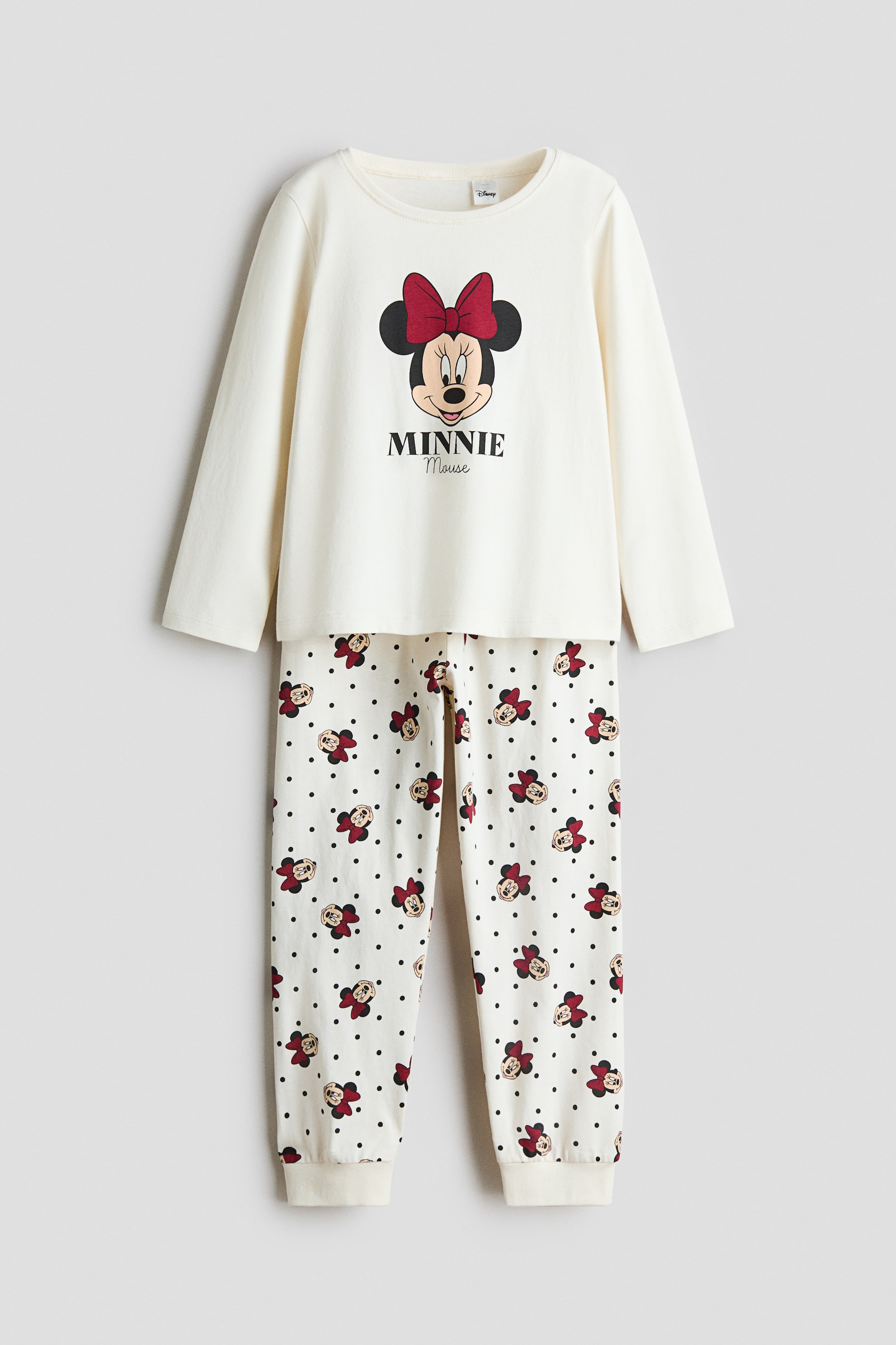 View larger image: A pyjama set is shown from the front, featuring an off-white, long-sleeved top with a round neckline and a large Minnie Mouse graphic printed on the chest. The matching cream-coloured pants feature an all-over pattern of smaller Minnie Mouse prints and black polka dots, ending with ribbed ankle cuffs.