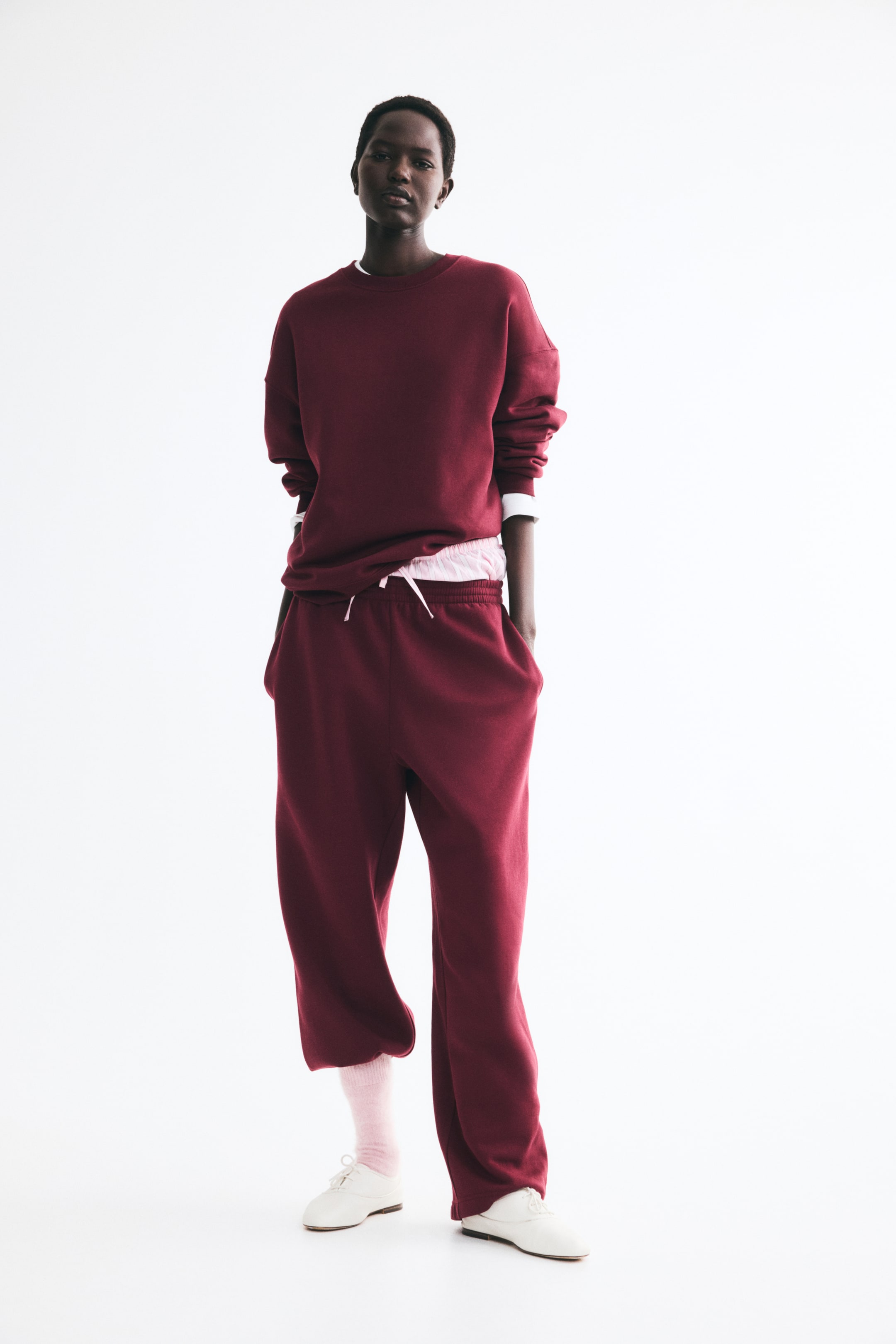 View larger image: The model is wearing burgundy sweatpants and standing with their hands in the side pockets. The straight-leg pants feature an elasticized waistband with a pink drawstring and are styled with a matching burgundy sweatshirt.