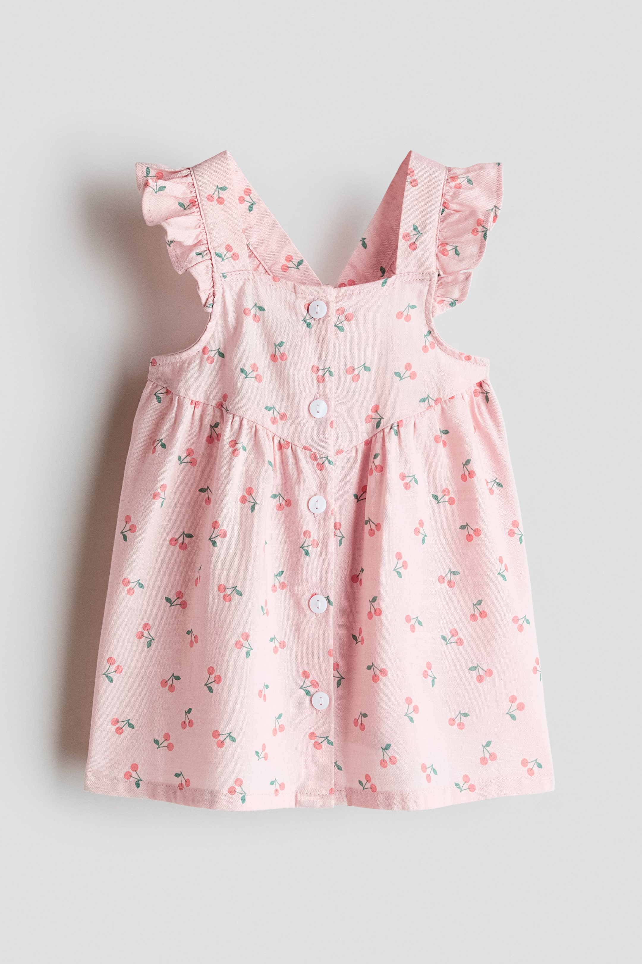 Ver imagen más grande: El vestido sin mangas de largo hasta la rodilla se presenta de frente, mostrando un tejido de algodón aireado con un estampado de cerezas. Cuenta con tirantes adornados con volantes, un escote cuadrado, botones blancos que recorren la parte delantera y una costura fruncida en la cintura.