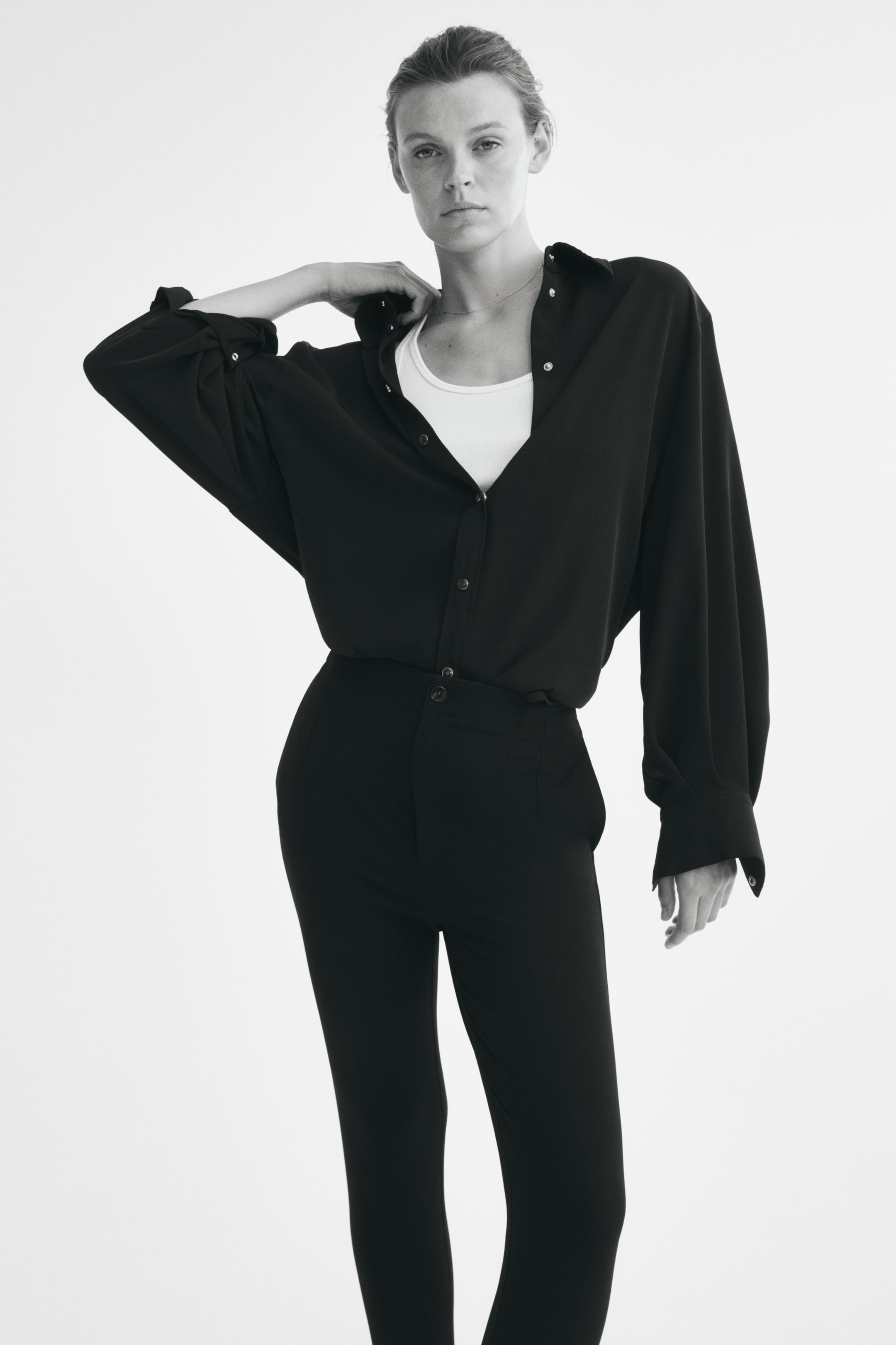 View larger image: The model is wearing fitted, high-waisted black trousers featuring a button fastening and front darts. The trousers are styled with a tucked-in open black button-up shirt layered over a white vest top.