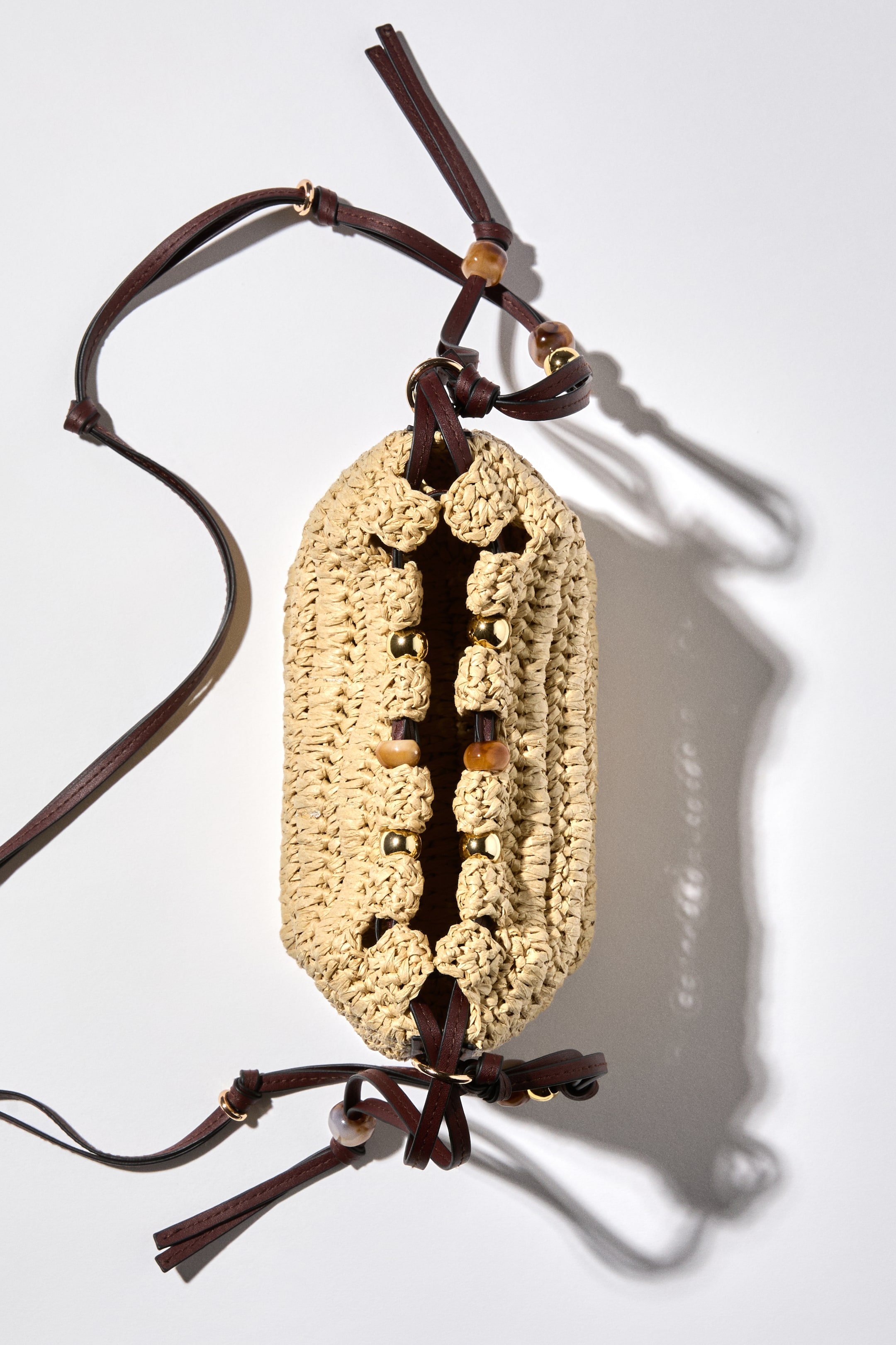 View larger image: A crossbody bag featuring a braided paper straw construction and a drawstring closure at the top. Its long, two-stranded shoulder strap and the drawstring are both detailed with knotted elements and plastic beads in various designs.