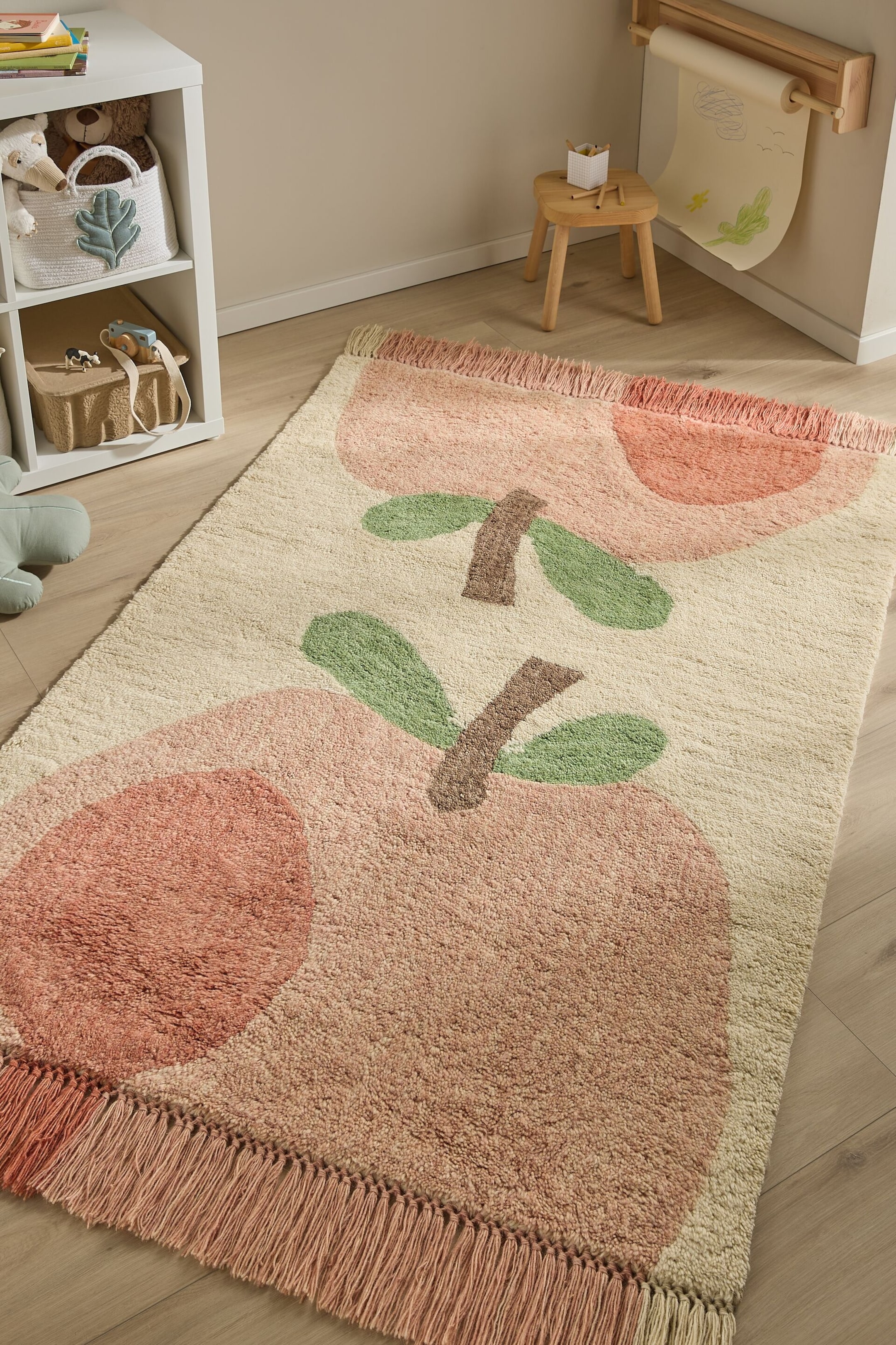 Ver imagem maior: O tapete apresenta grandes designs abstratos de maçãs em tons de rosa e pêssego, com caules castanhos e folhas verdes sobre um fundo branco sujo. Exibe uma textura felpuda e bordas com franjas rosa a condizer, estendido plano.