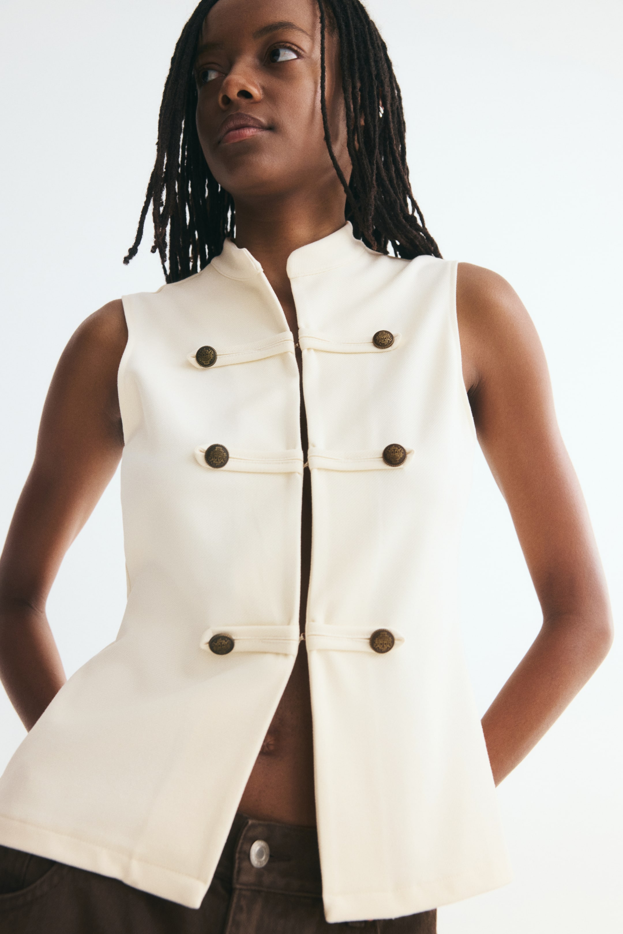 Visualizza l'immagine ingrandita: Un gilet aderente color crema è mostrato frontalmente, mettendo in risalto un colletto alla coreana e un design senza maniche. Il gilet presenta delle linguette orizzontali decorative con bottoni metallici incisi lungo la parte frontale e si chiude con chiusure nascoste sopra un orlo dritto con spacchetto.