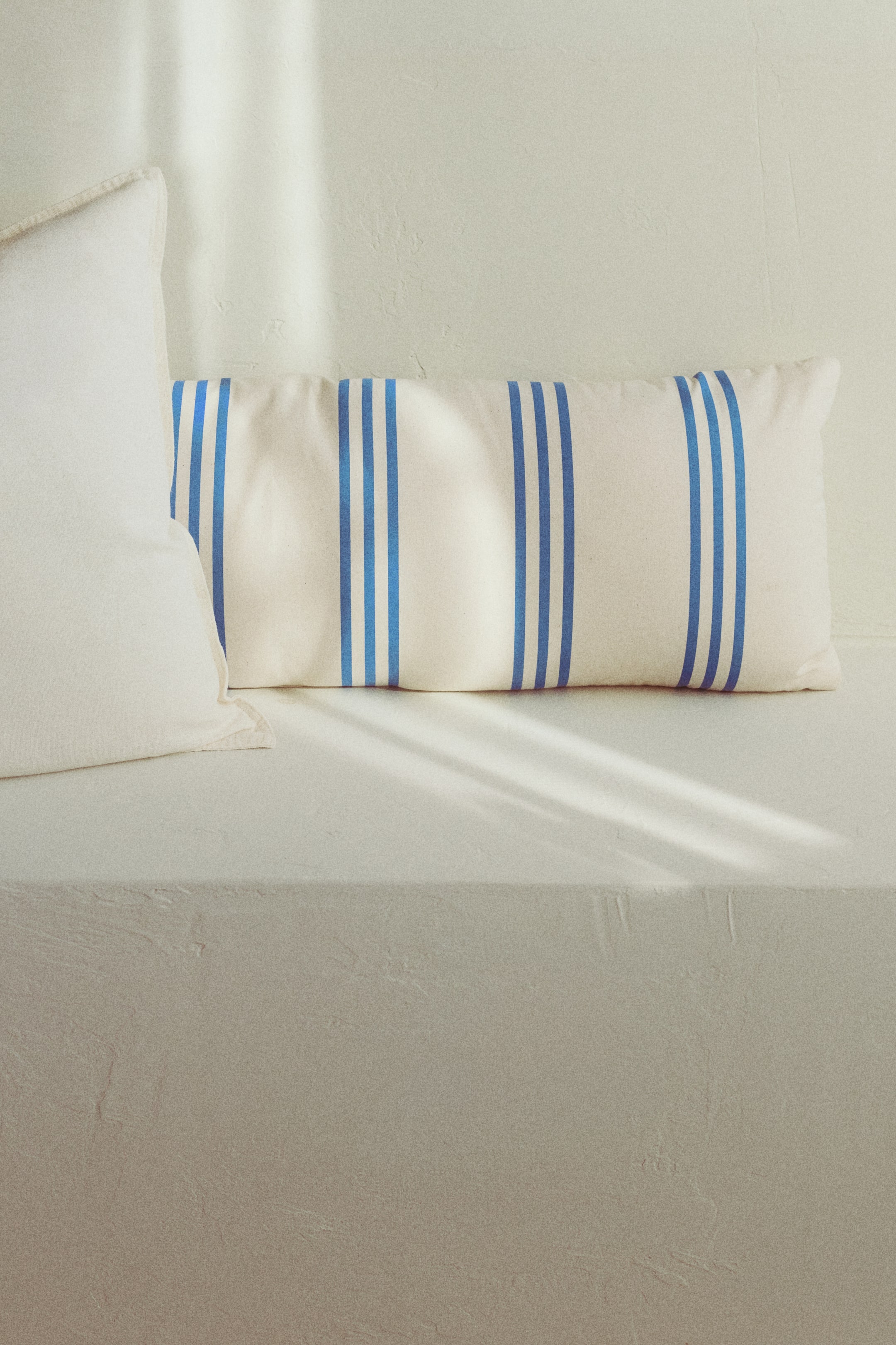 Ampliar la imagen: Un cojín rectangular se presenta de frente, con franjas triples verticales azules sobre un lienzo de algodón color blanco roto repelente al agua, adecuado para uso en exteriores.