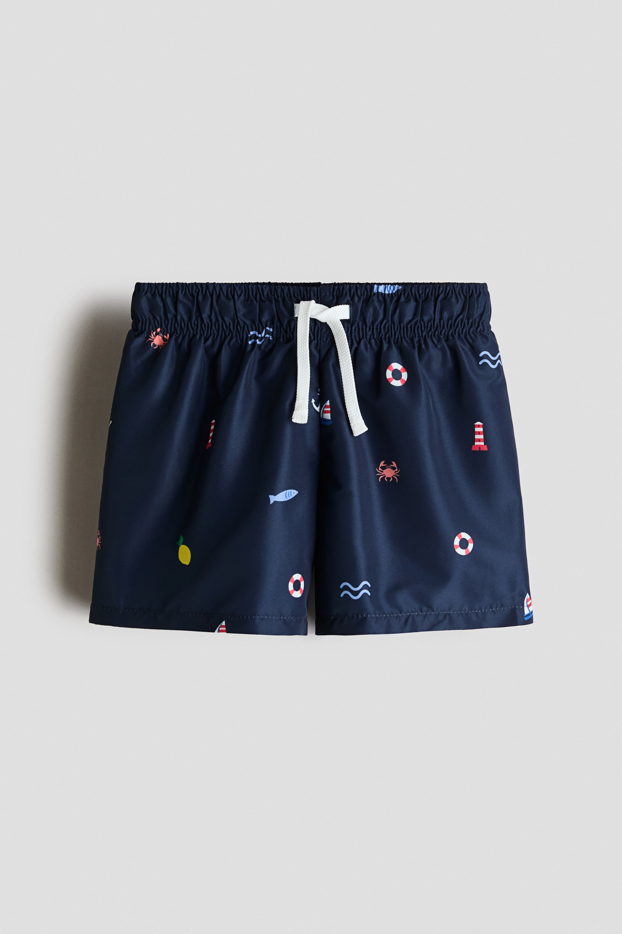 Grotere afbeelding bekijken: Gedrukte zwembroek wordt aan de voorkant getoond, met een marineblauwe basiskleur en een veelkleurig nautisch patroon. Ze hebben een elastische tailleband met een witte trekkoord in een strik geknoopt.
