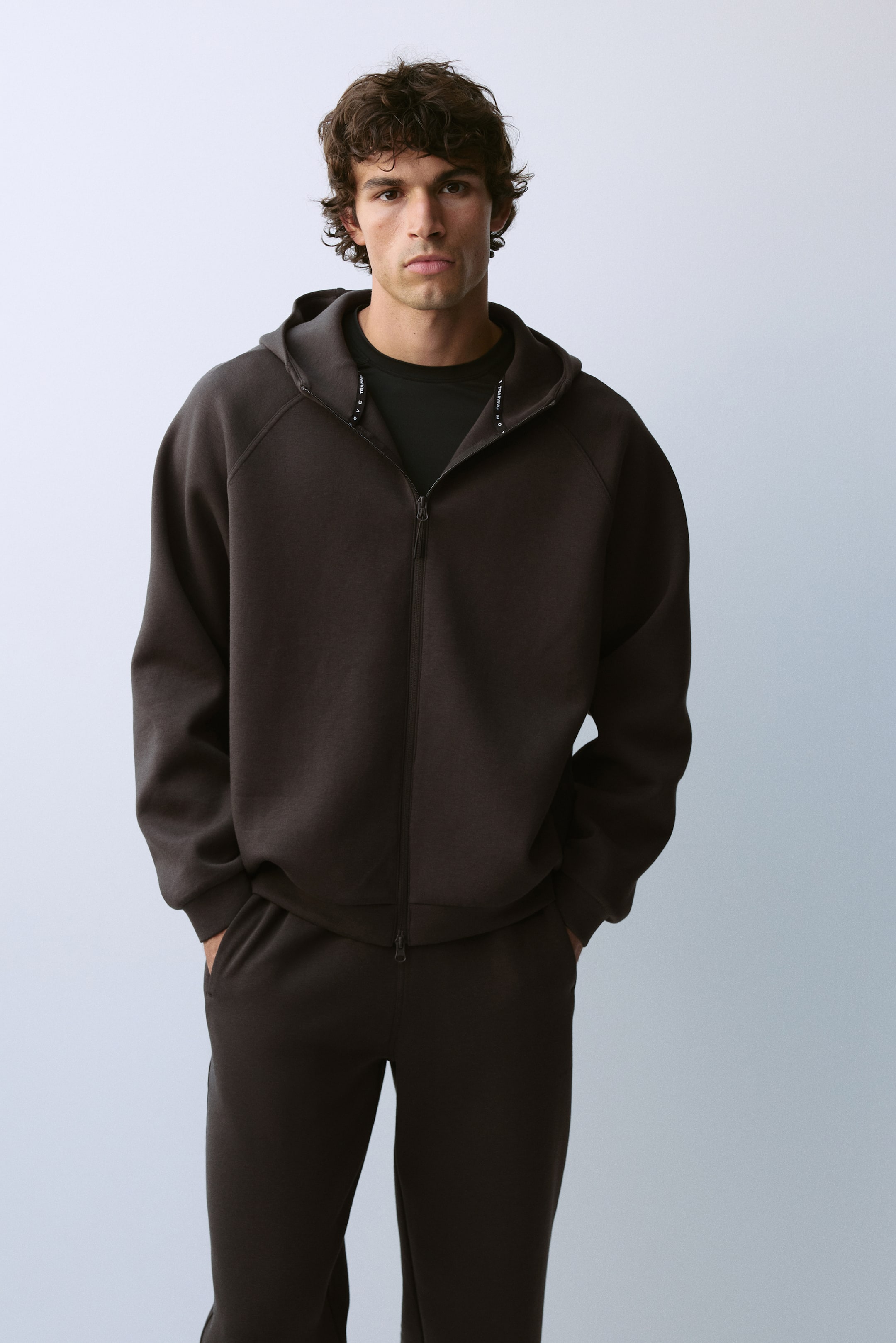 View larger image: A dark brown zip-up hoodie featuring raglan sleeves and a hood is being worn by the model over a black crew-neck T-shirt. The hoodie is paired with matching dark brown sweatpants, with the model resting their hands in the pockets.