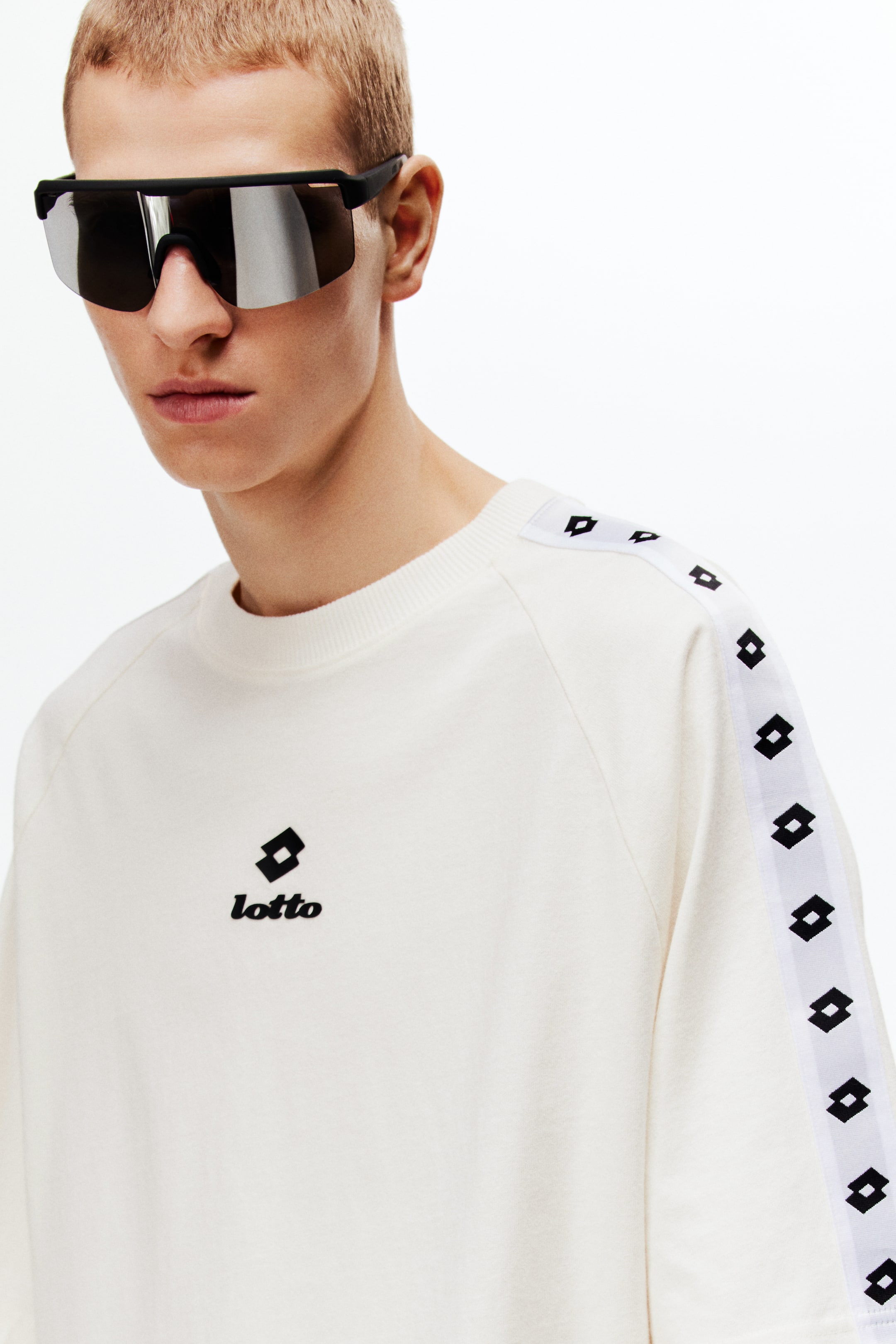 Ampliar la imagen: La modelo lleva puesta una camiseta de color blanco roto confeccionada en jersey de algodón de gran peso, que presenta un estampado del logo "lotto" en negro en el frente. Cuenta con un cuello redondo con ribete y mangas raglán cortas que muestran una cinta tejida con un patrón geométrico negro repetido a lo largo de los lados, ofreciendo un ajuste holgado.