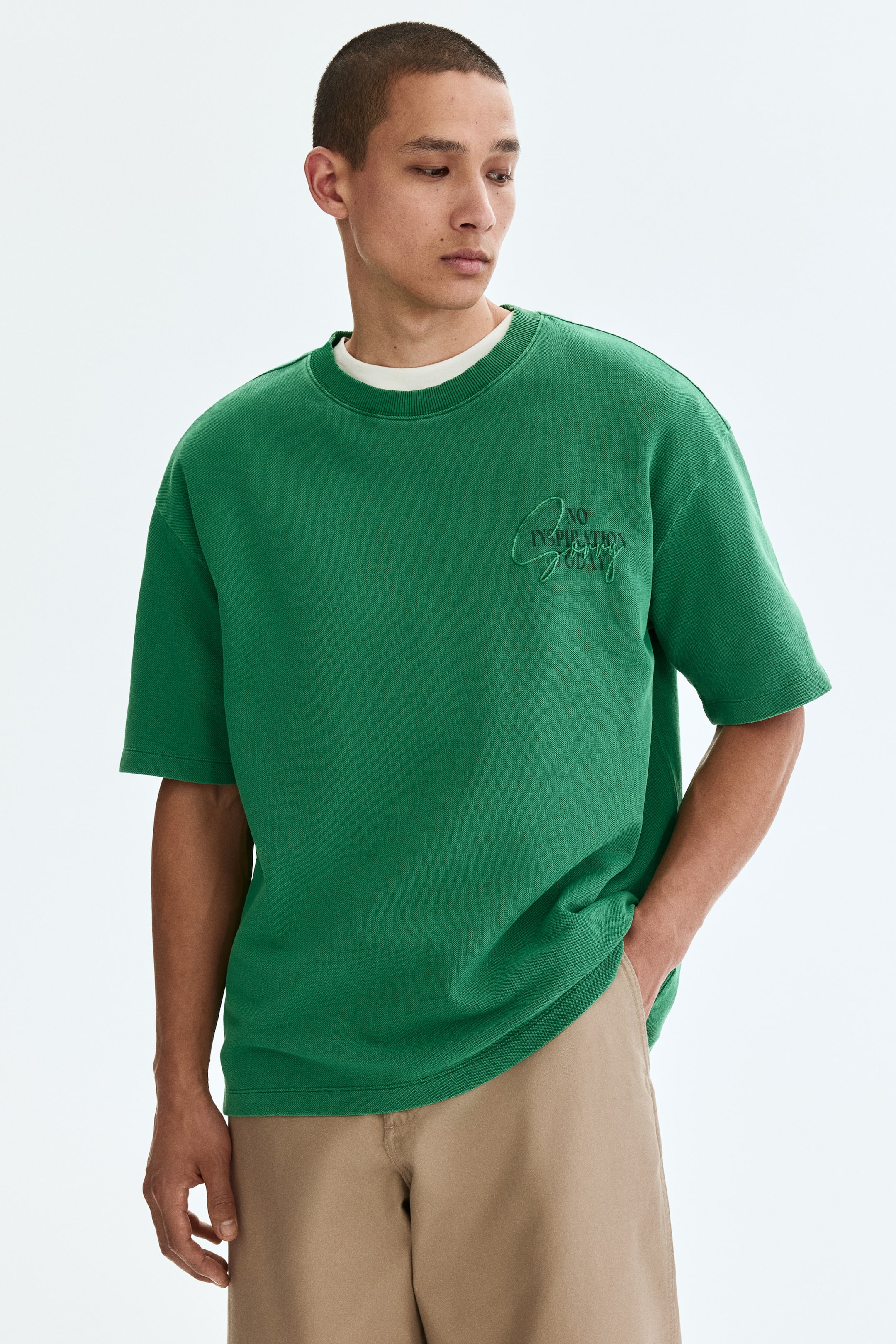 View larger image: The model is donning a green T-shirt crafted from heavyweight cotton jersey, showcasing a round neckline with ribbed trim and an embroidered motif that reads 'NO INSPIRATION TODAY' on the front. The T-shirt's relaxed fit and straight-cut hem are evident, complemented by a white undershirt layered underneath. The model's hand is casually placed in the pocket of khaki trousers.