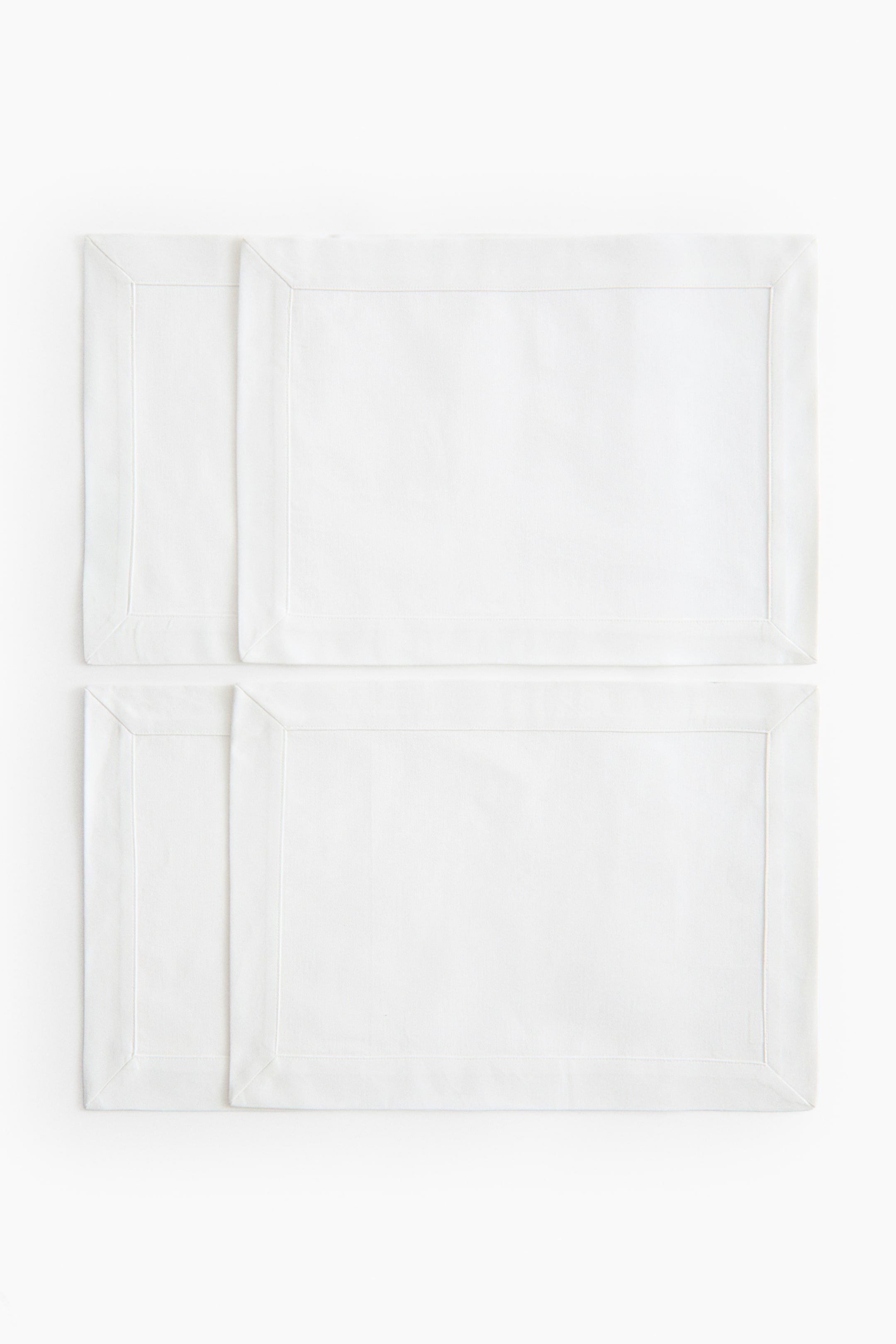 Agrandir l'image: Quatre sets de table rectangulaires blancs sont posés à plat, chacun présentant une large bordure cousue, créée par des ourlets repliés sur ses bords. Ils sont disposés en deux rangées, avec deux sets de table partiellement superposés dans chaque rangée.