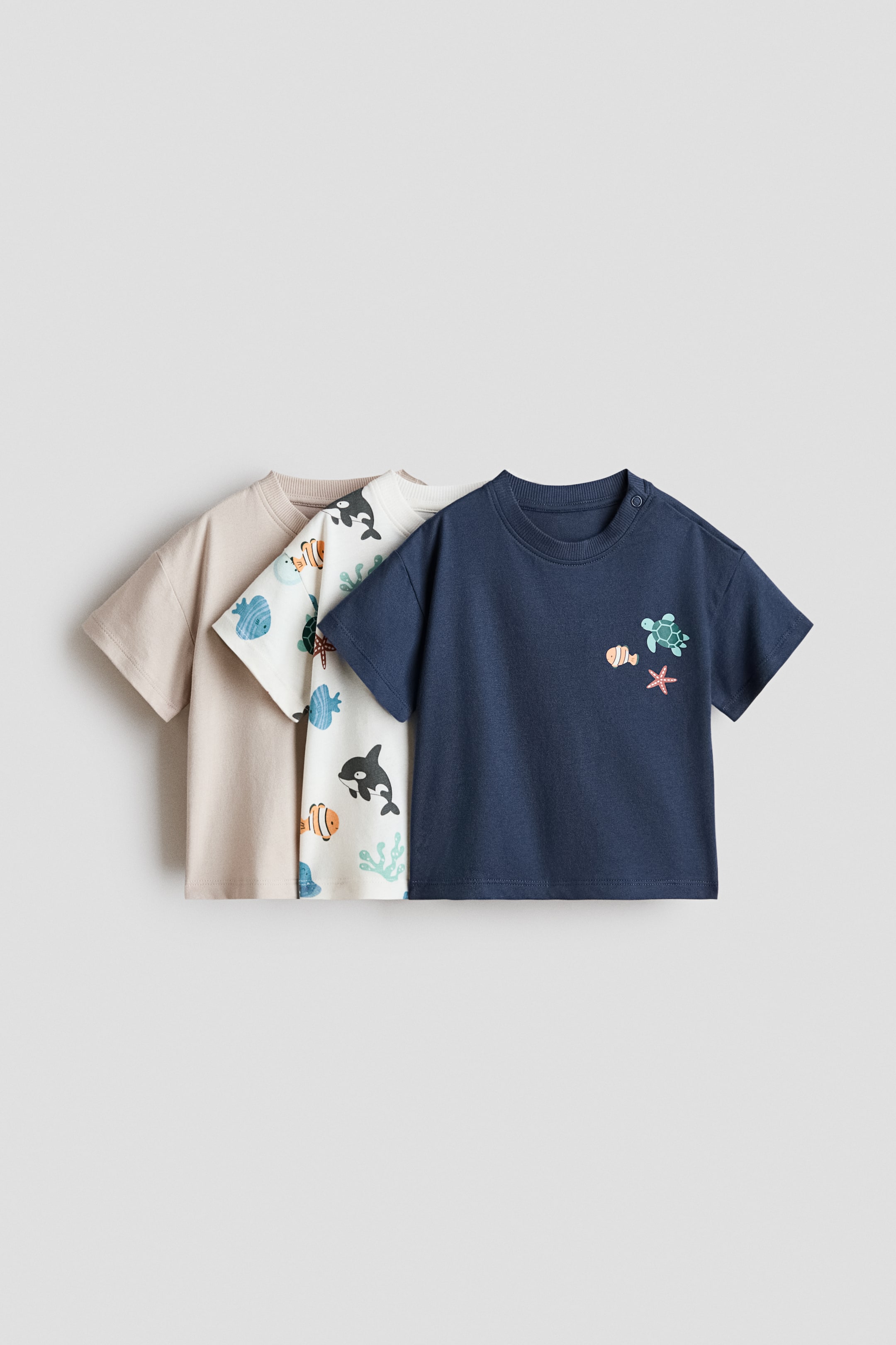 View larger image: Presenting a 3-pack of cotton T-shirts, each one layered to display its front. The front navy blue T-shirt showcases a small sea creature graphic and shoulder snap buttons, while the middle off-white T-shirt features an all-over sea creature pattern. The back T-shirt is plain beige.